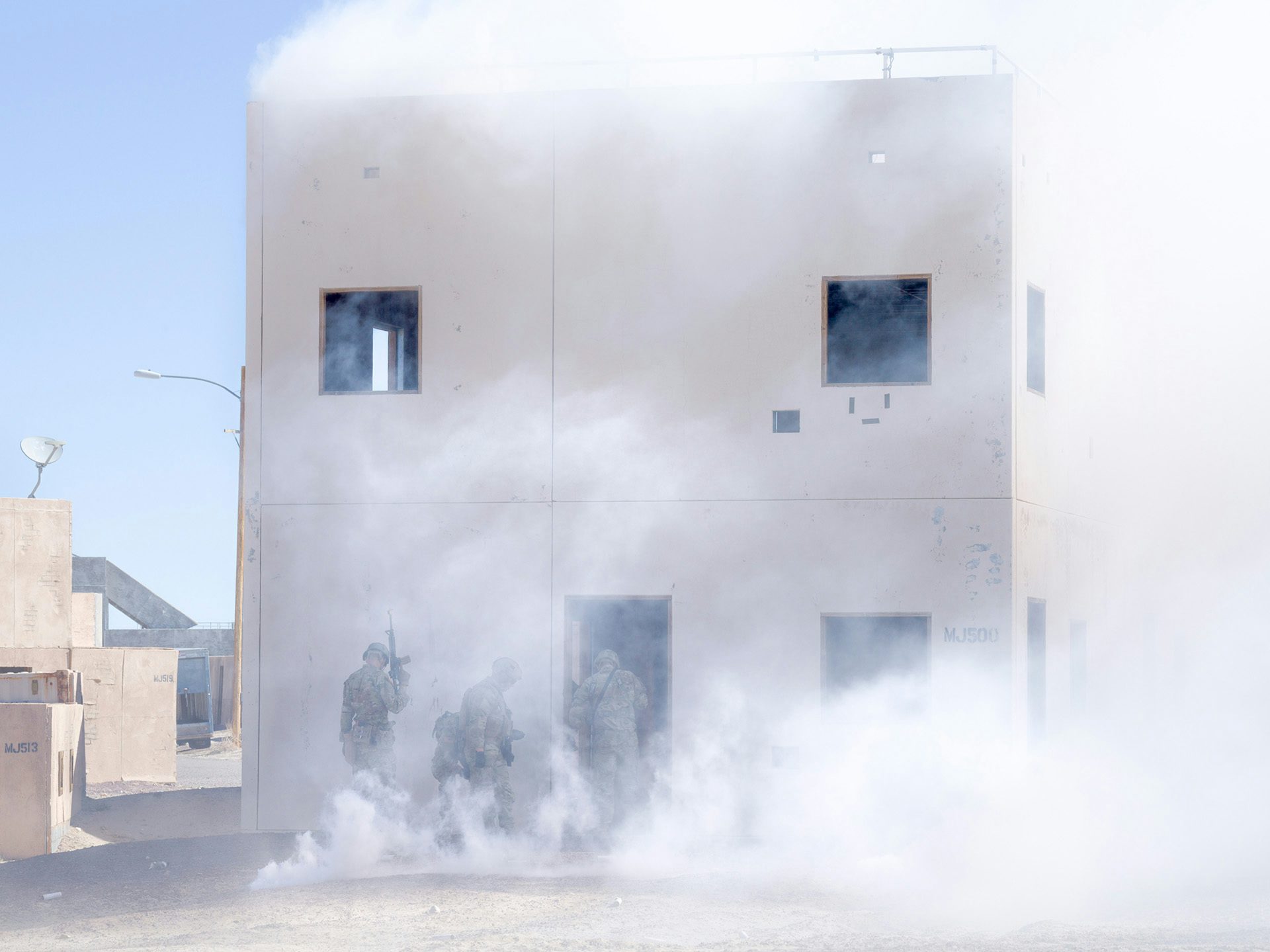 Image from American Glitch by Andrea Orejarena and Caleb Stein showing the frontage of a house in an Iraqi village simulation shrouded in smoke
