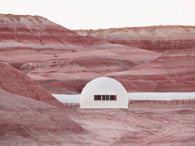 Image from American Glitch by Andrea Orejarena and Caleb Stein showing a Mars simulation, featuring a white semi circular building surrounded by dusty red-brown hills