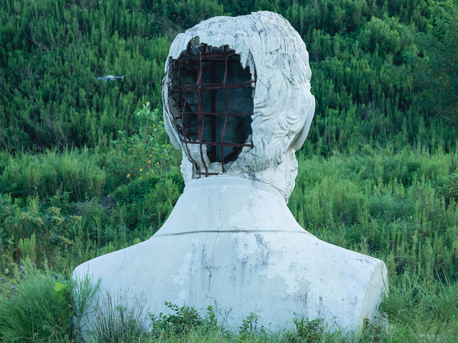 Image from American Glitch by Andrea Orejarena and Caleb Stein showing the back of a large statue's head crumbling away surrounded by trees