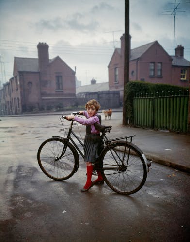 Photograph by Evelyn Hofer from 1966 showing a young girl wearing a grey skirt and knee high red socks, who is tiptoeing over an adult sized bicycle frame in a residential street in Dublin