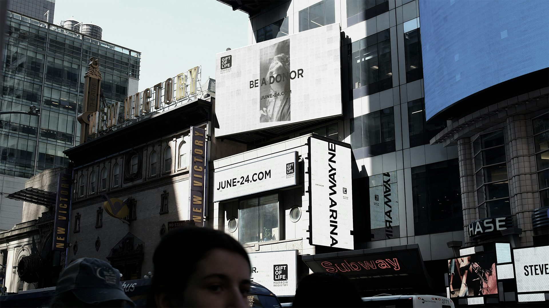 Photograph of white billboards in Times Square, which read the names Wayne and Marina joined by the letter 'M' and 'W', and another that reads 'Be a donor', and another containing a URL