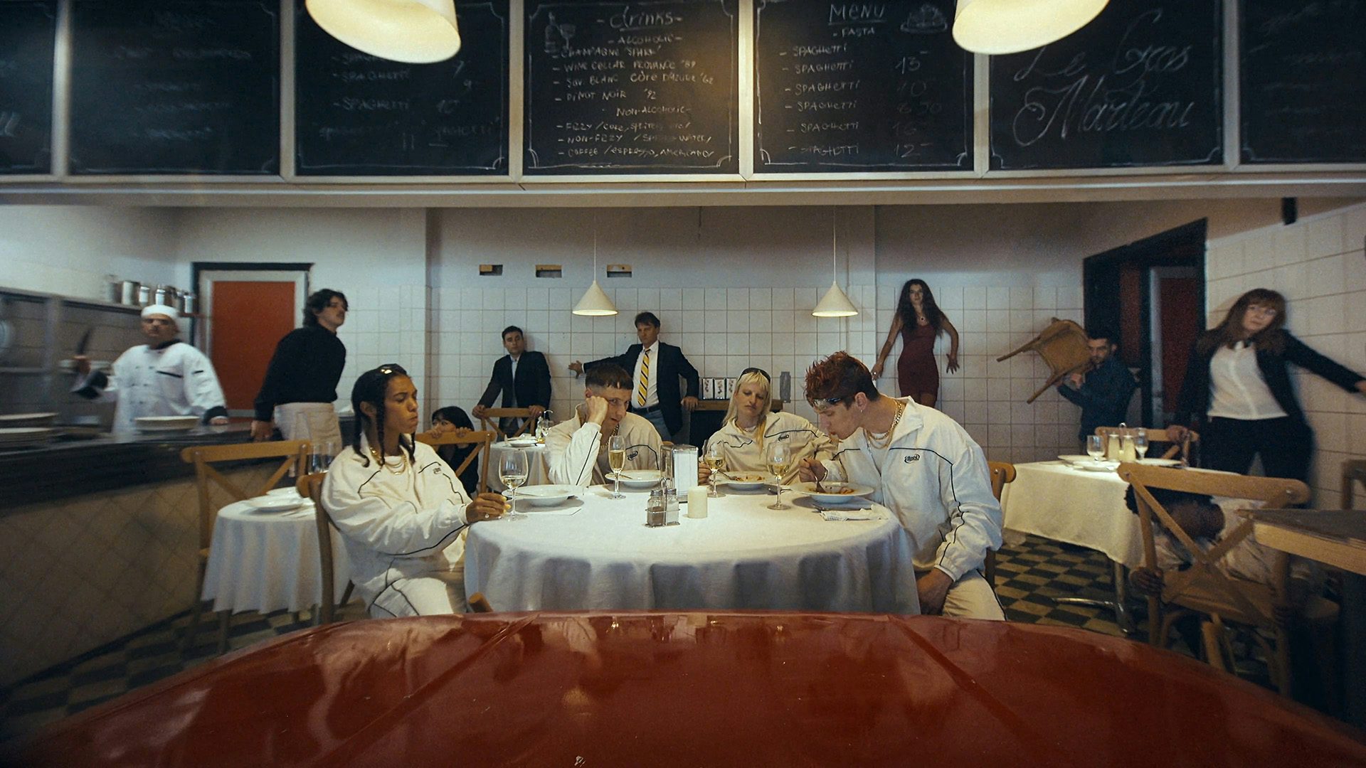 Still from Oscar Hudson's music video for Big Hammer by James Blake showing four young people wearing matching white tracksuits and eating food in a restaurant, which is lined with scared looking restaurant staff and guests