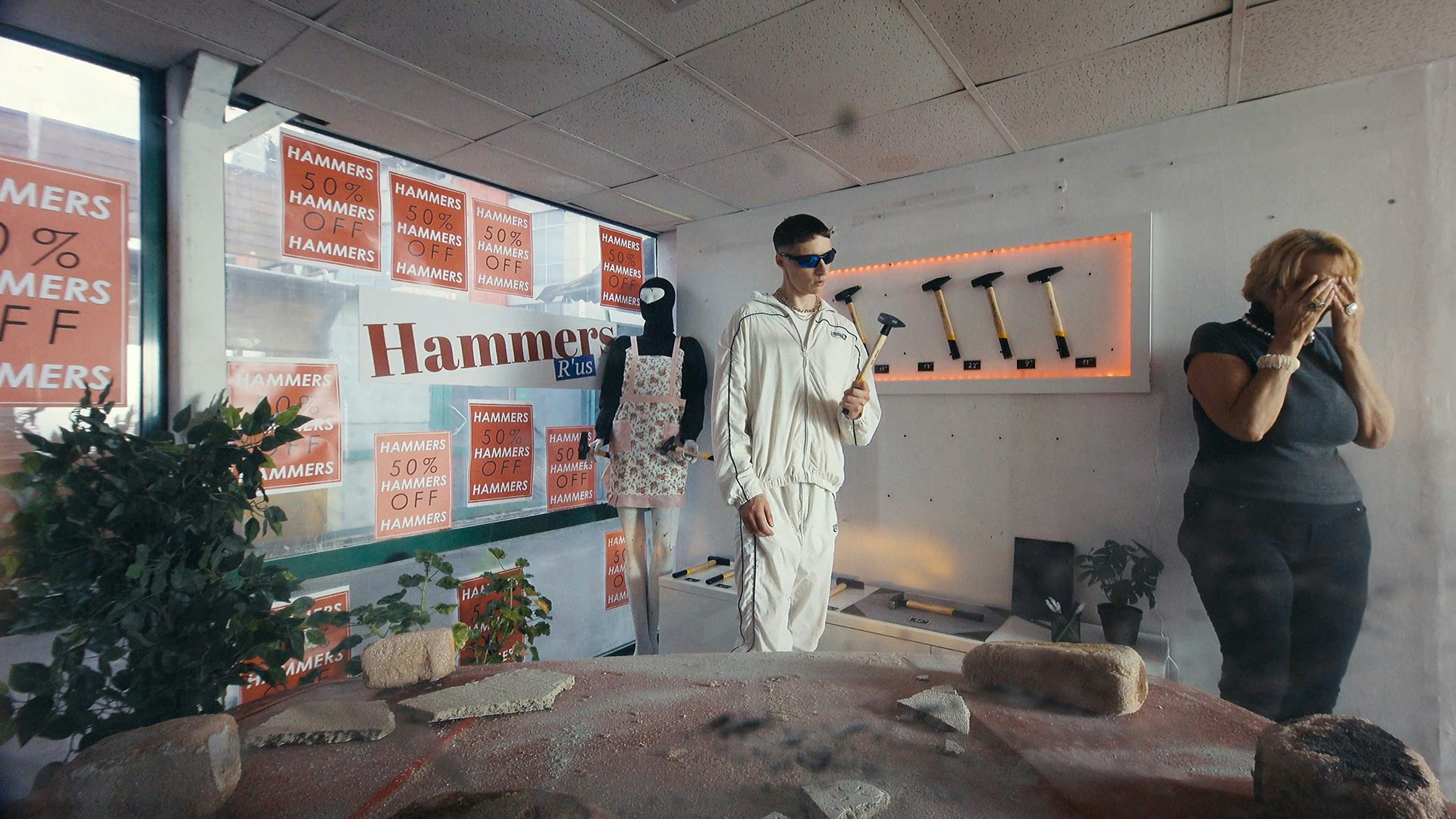 Still from Oscar Hudson's music video for Big Hammer by James Blake showing a young person wearing a white tracksuit and sunglasses, holding a hammer in a hammer store with a shop assistant covering their eyes in the corner