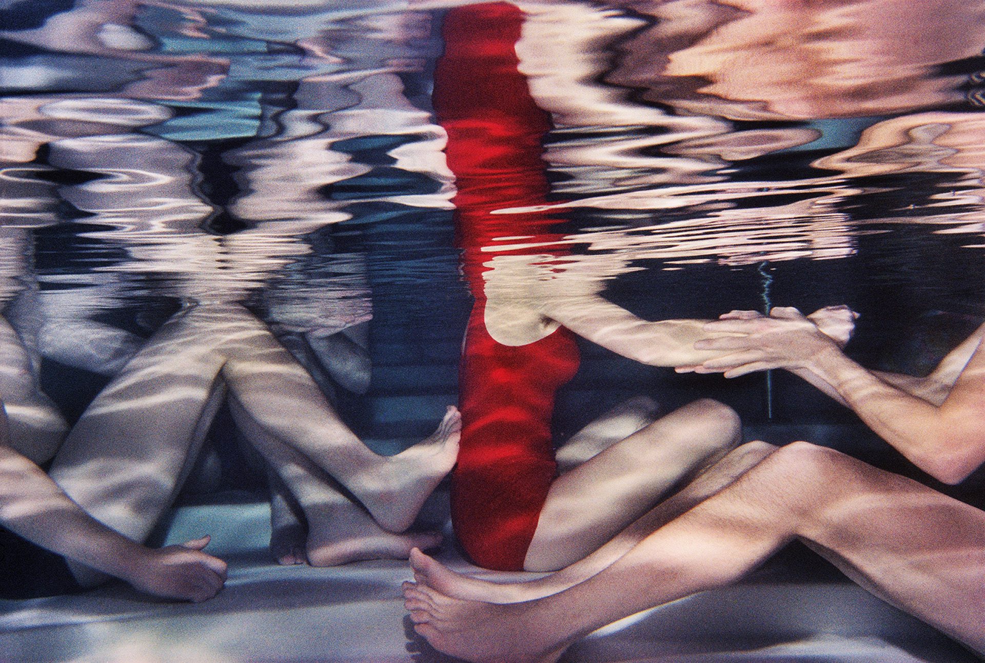 Photograph by Larry Sultan from his series Swimmers showing rows of people sat on the bottom of a swimming pool including a person wearing a bright red swimming costume