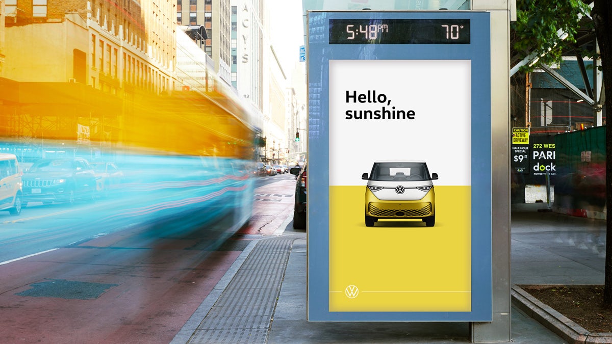 Photo of a vertical VW ID Buzz poster in a busy city street, with the tagline 'Hello sunshine' above a head-on image of the VW bus in white and yellow, with a matching white and yellow background