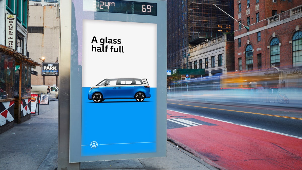 Photo of a vertical poster in a busy city street created for VW's ID Buzz campaign, showing the tagline 'A glass half full' above an image of a blue and white VW bus with a matching blue and white background