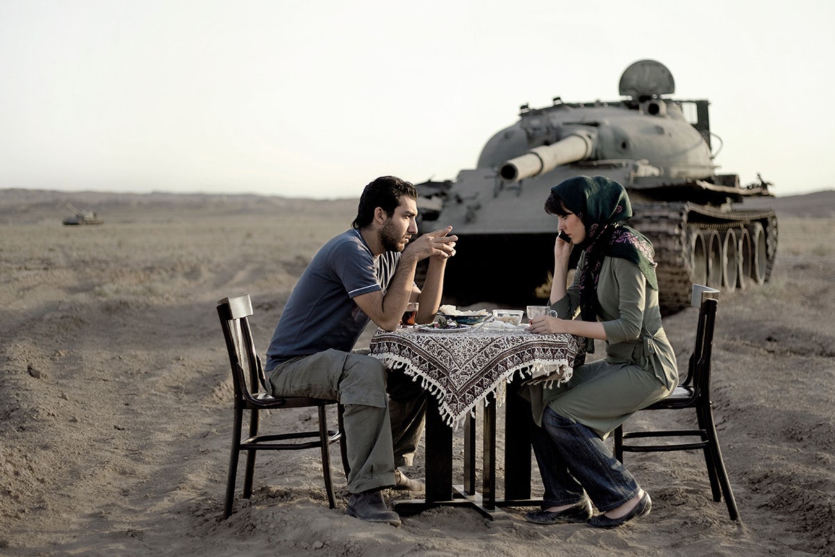 Photograph by Gohar Dashti showing a couple sat at a table drinking and eating, which has been positioned in a desert landscape with a tank directly behind them
