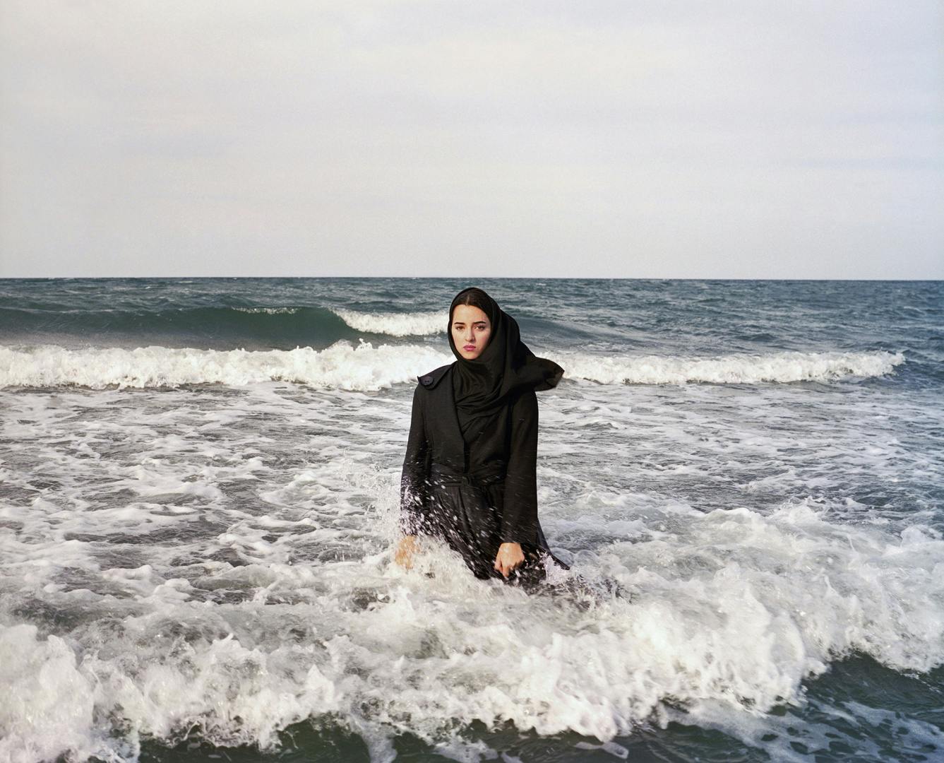 Photograph by Newsha Tavakolian showing a young woman wearing hijab and dark attire stood in the sea at the shore