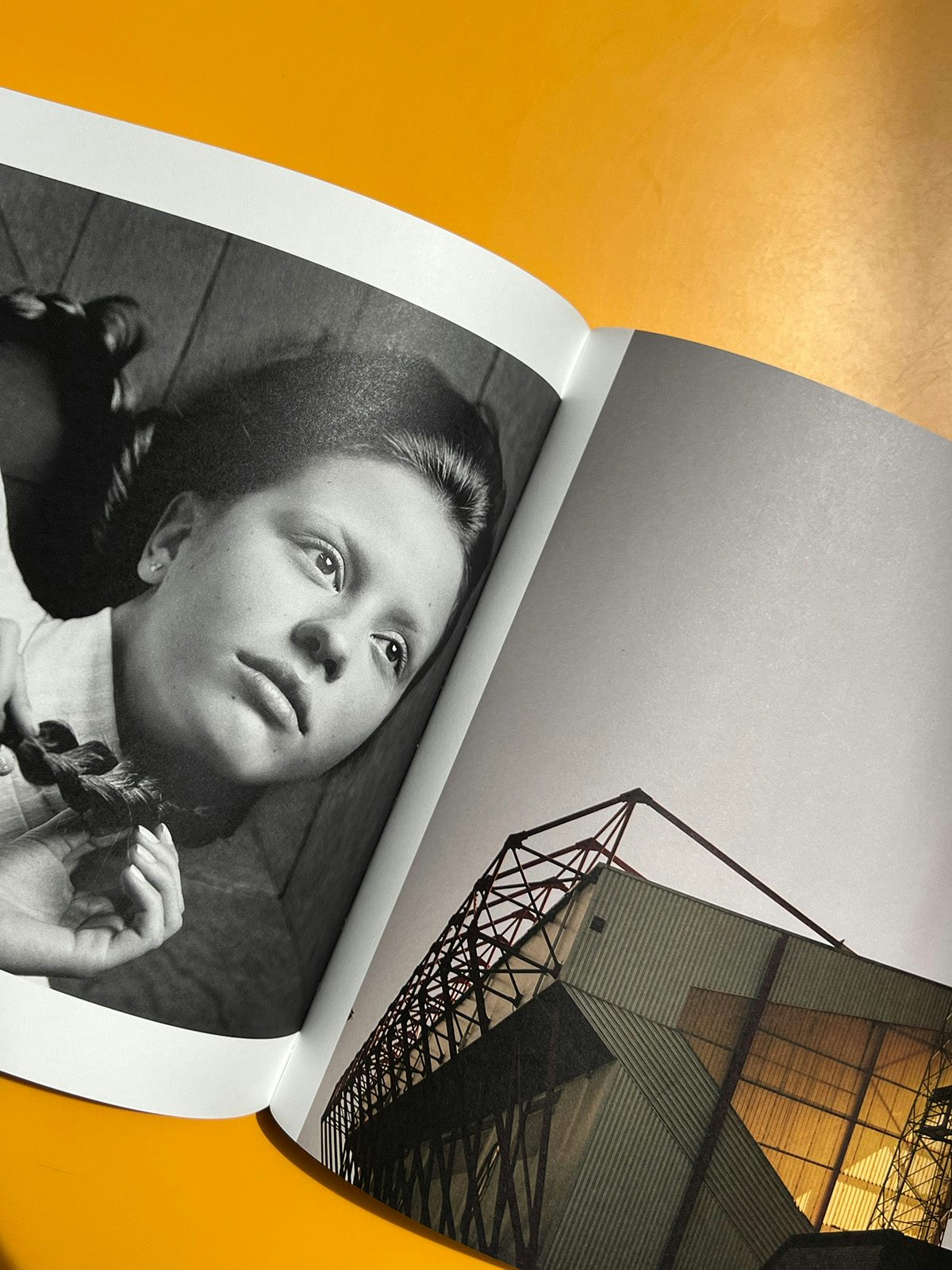 Photo of Alasdair McLellan's Home and Away photo books on an orange tabletop, featuring a black and white portrait of Mia Goth with plaits on the left hand page and an exterior photo of a stadium on the right hand page