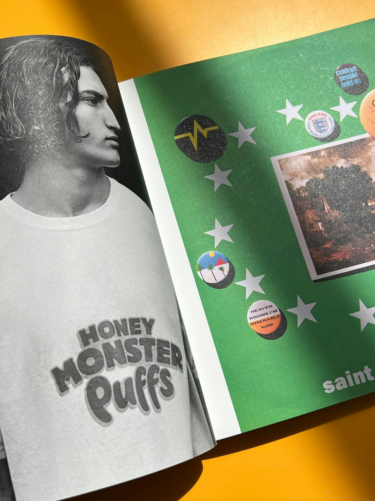 Photo of Alasdair McLellan's Home and Away photo books on an orange tabletop, showing a black and white portrait of a person with curly chin-length hair wearing a t-shirt that reads 'Honey Monster Puffs', next to another photograph of badges on a green background