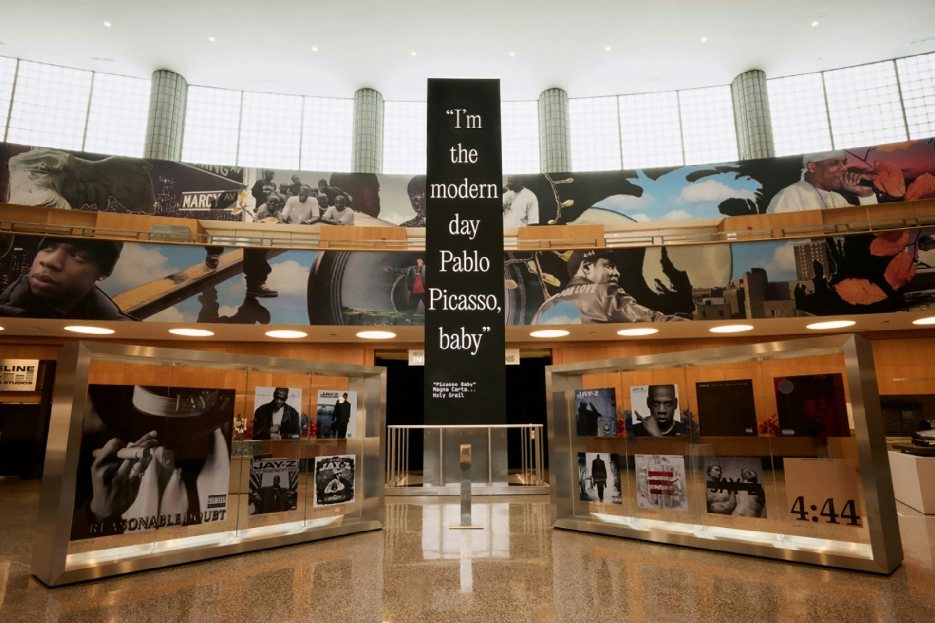 Photo of the main hall at the Brooklyn Public Library showing Jazz Grant's mural of Jay Z lining the space, and a vertical banner that reads 'I'm the modern day Pablo Picasso, baby'