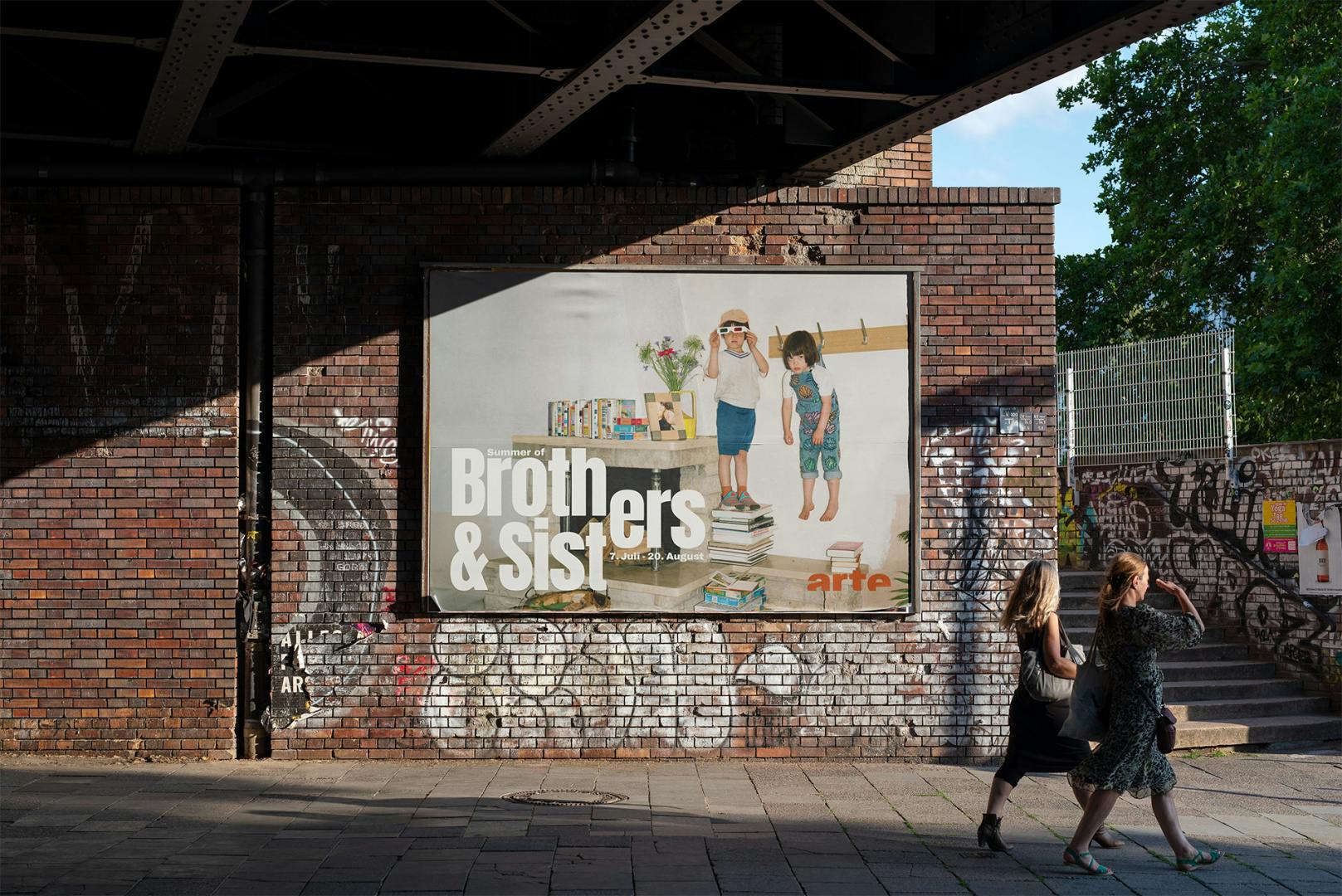 Photo of Arte's Brothers and Sisters campaign on a brick wall. The poster shows a photo of a child wearing sunglasses standing on a tower of books with another child with a fringe and dungarees hanging from a coat hook. The letters 'er' are split between the words 'Brothers' and 'Sisters'
