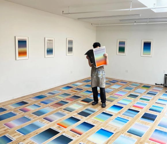 Photograph of artist Sho Shibuya holding one of his sunrise artworks painted on a New York Times cover concealing his face, while standing on a glass floor with New York Times covers under foot