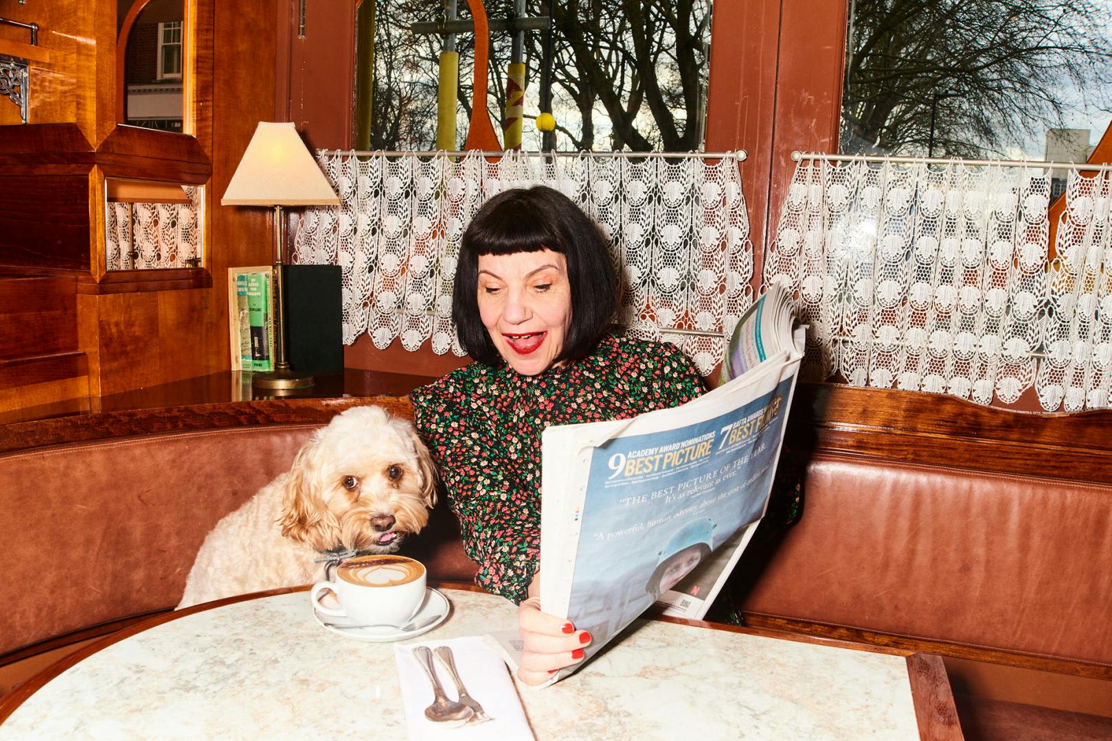 Image from the Wolseley's new digital identity, shown in a photo of a person with a black bob and fringe sat at a table while holding a newspaper next to a small dog leaning over a coffee