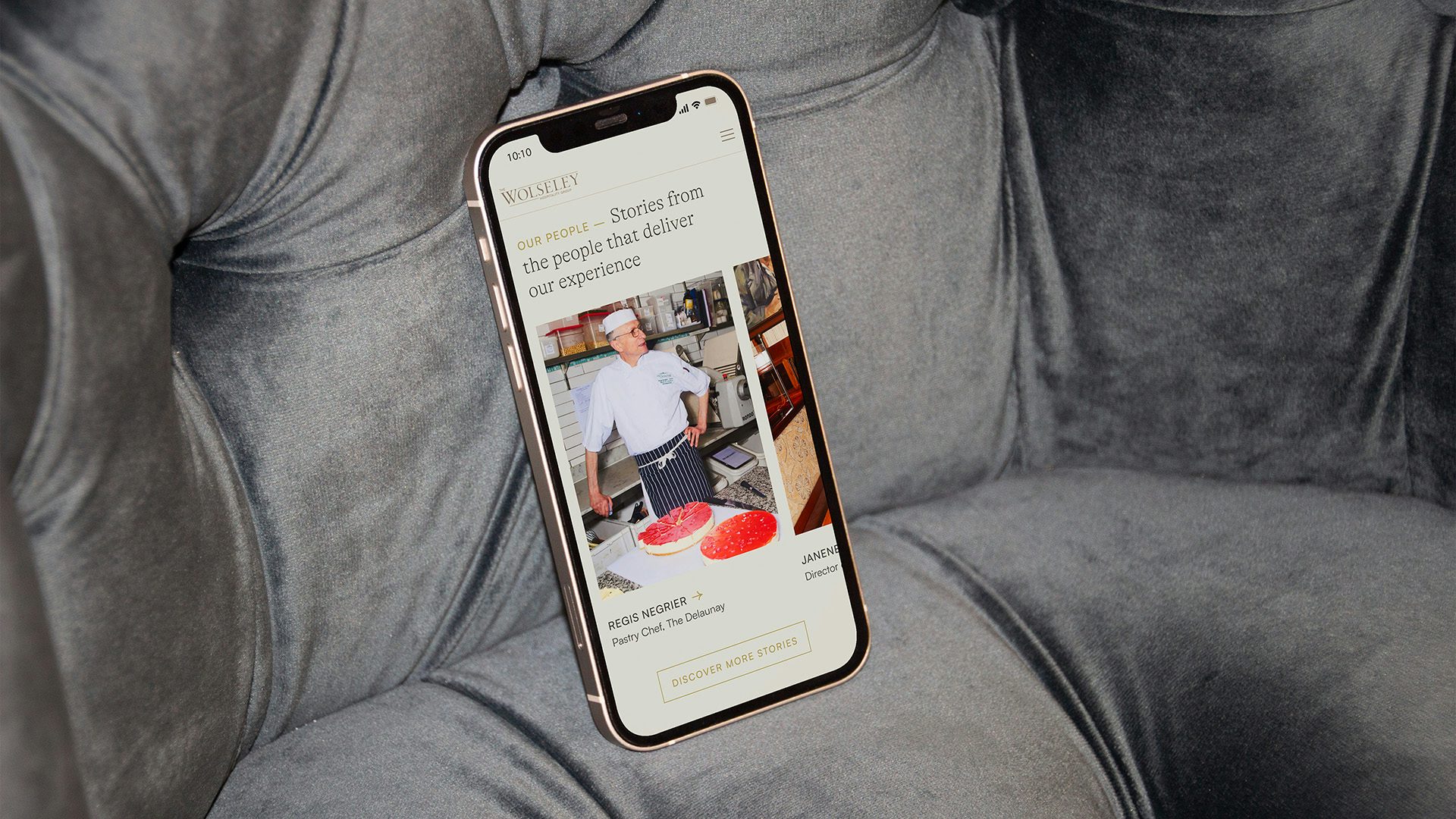 Image from the Wolseley's new digital identity, shown on a photo of a chef with a hand on their hip stood over a dessert and the page headline 'stories from the people that deliver our experience'. The web page is shown on an iPhone resting on a grey velvet couch