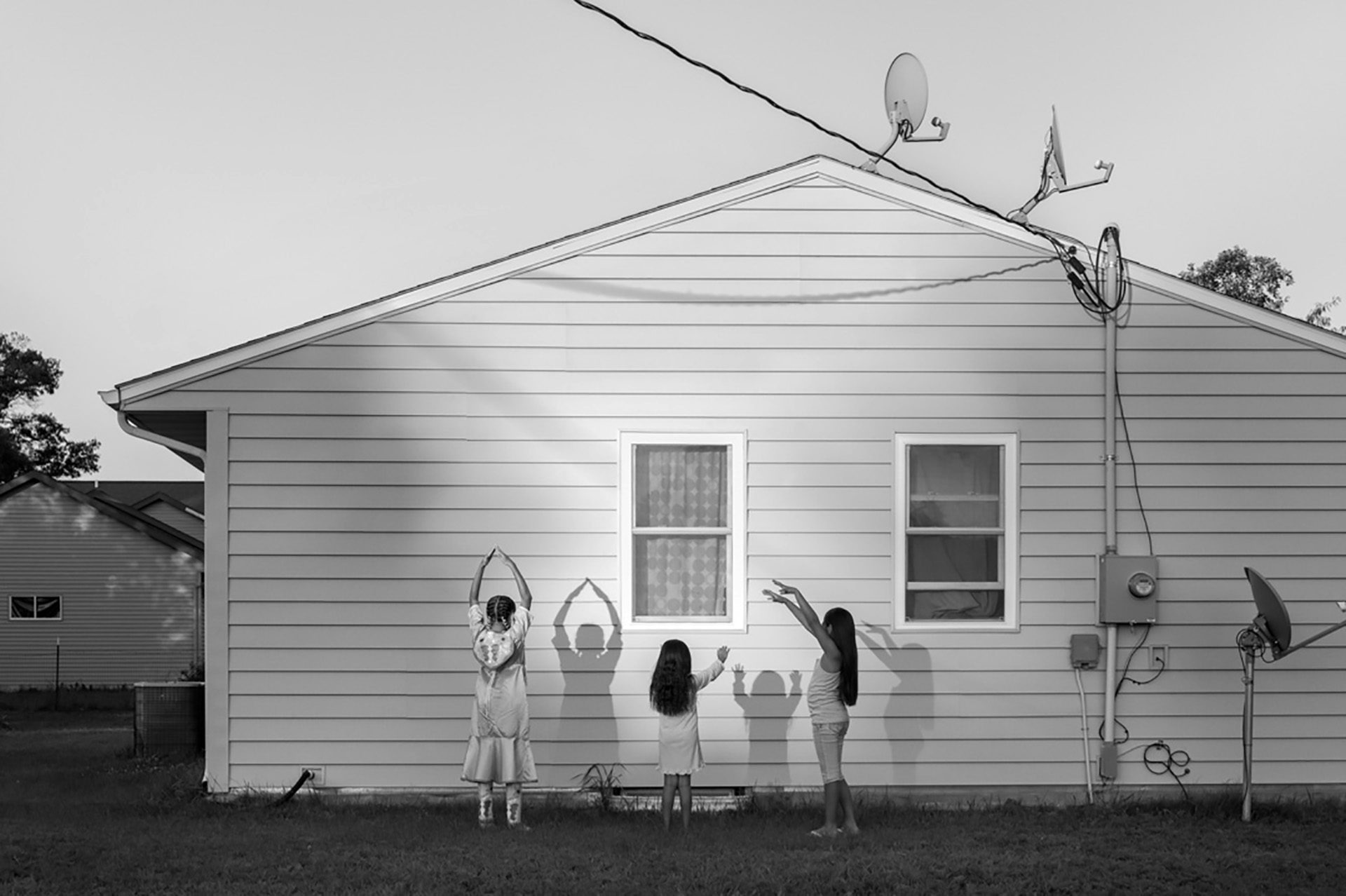 Photograph by Alessandra Sanguinetti of three girls facing a wood panelled house posing with their arms in the air and casting shadows onto the side of the building