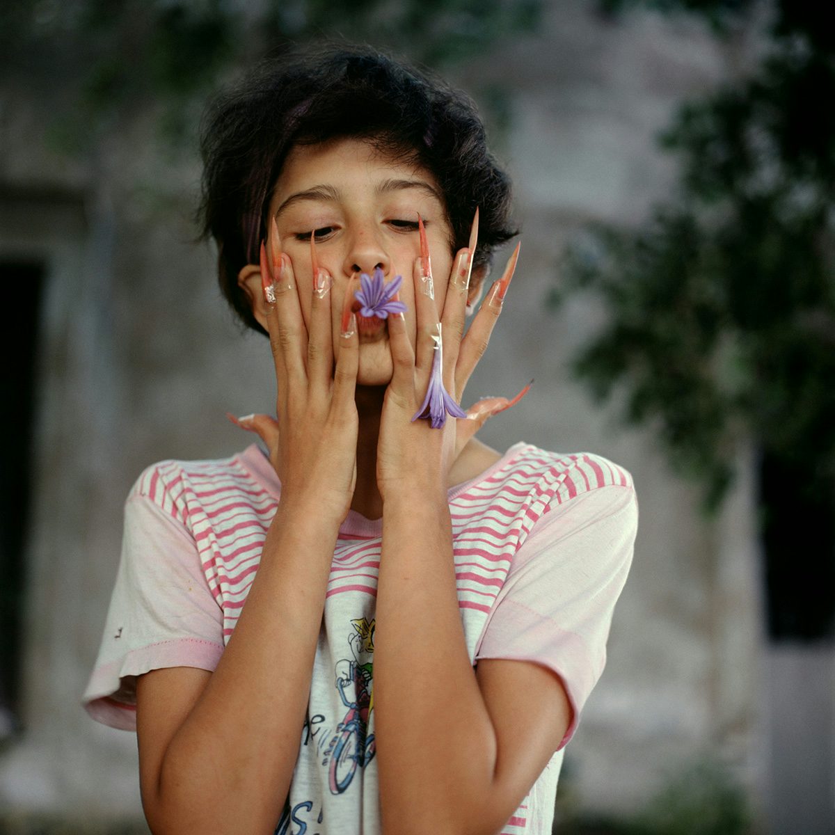 Photograph by Alessandra Sanguinetti of a person with dark cropped hair wearing a striped t-shirt and very long pointed nails, holding a purple petal to their lips