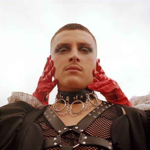 Photo by Dean Davies and student Kaye Lowe of a person wearing a leather and mesh chest brace, black puff sleeves, and a silver and black choker neck piece, with another person's hands in red gloves holding their face