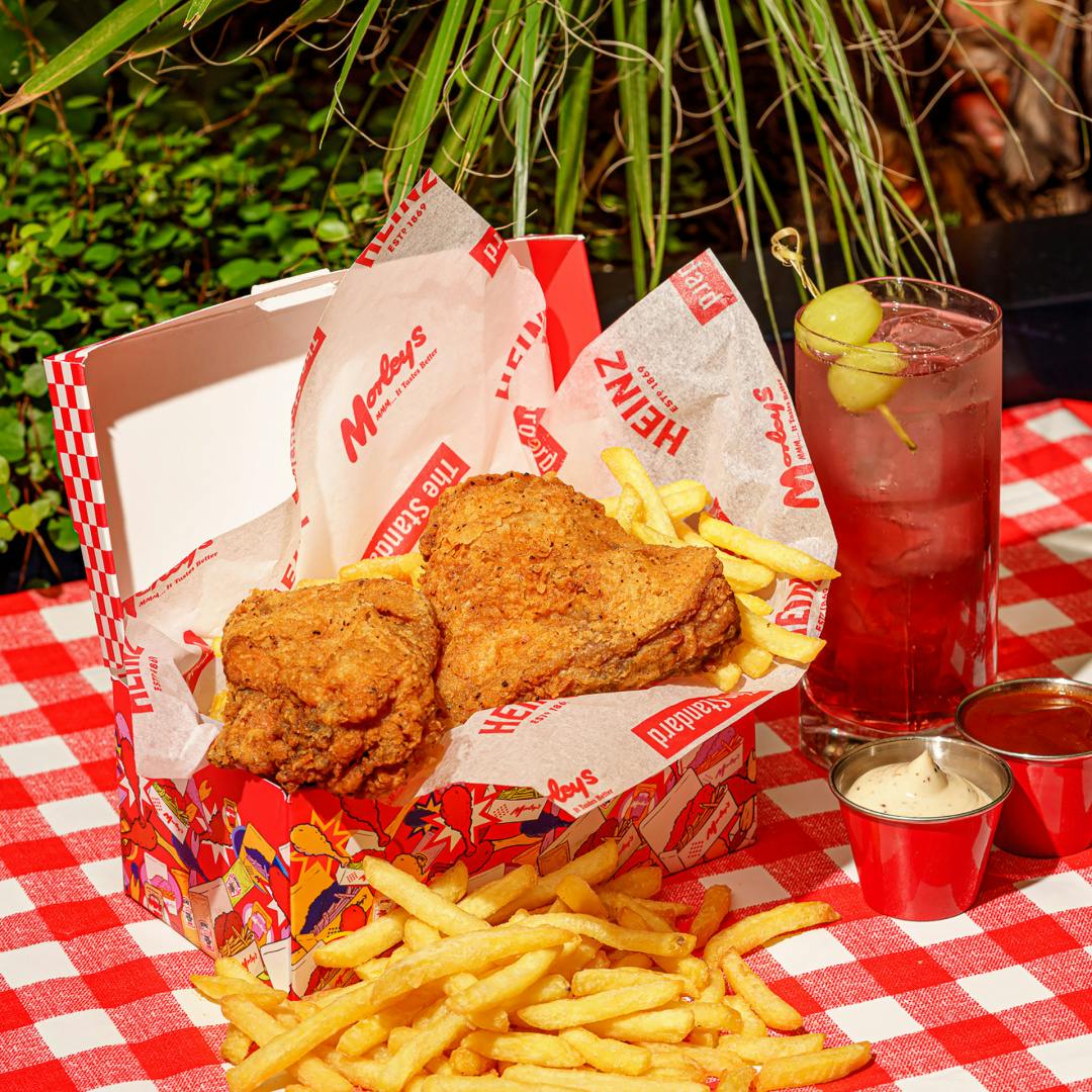 Photograph showing a box of fried chicken, a red drink with olives on a cocktail stick, dipping pots of sauce, and a stack of chips laid on a red gingham tablecloth