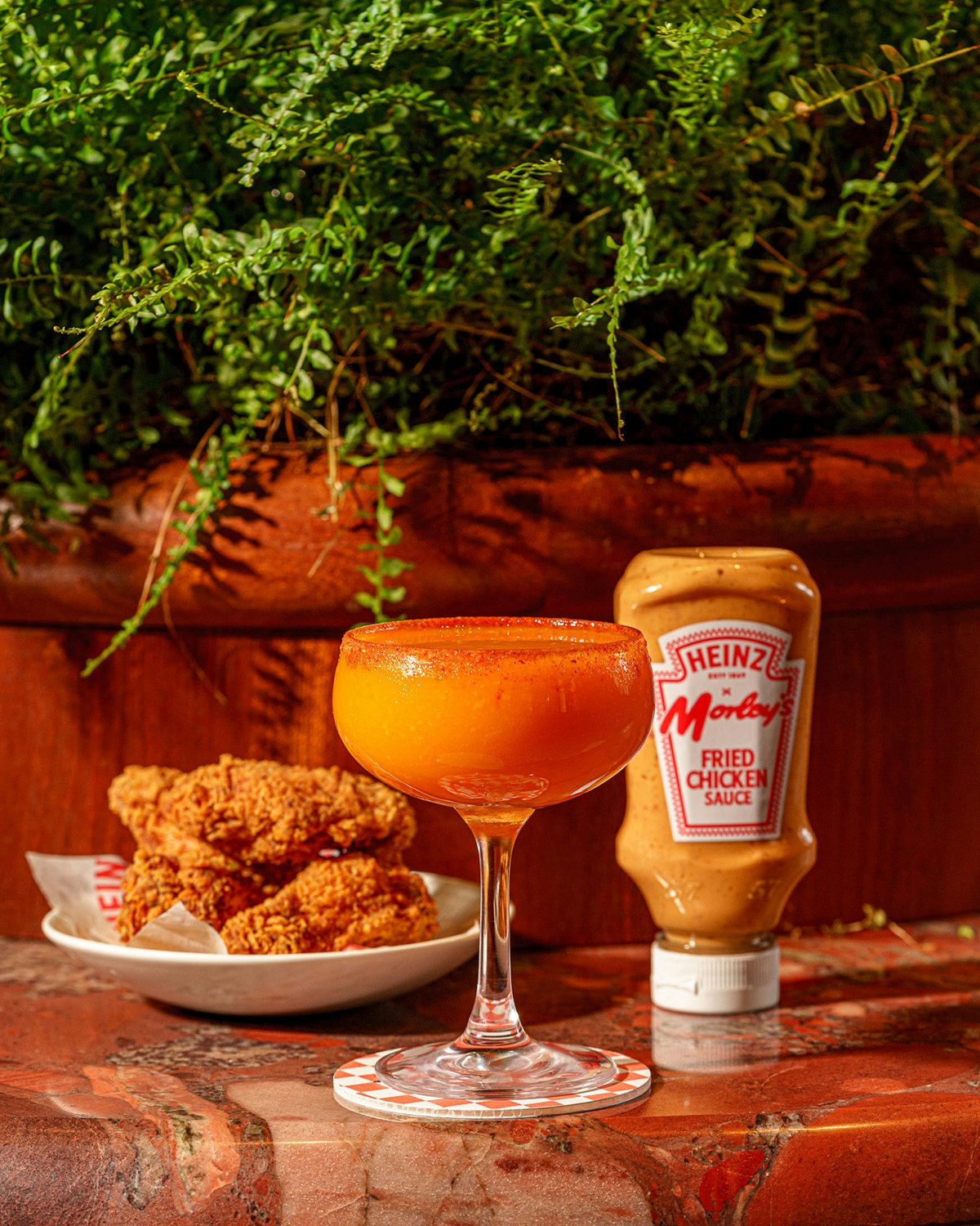Photograph showing an orange drnik in a cocktail glass, a plate of fried chicken, and a squeezy bottle of Heinz and Morley's Fried Chciken Sauce, arranged on a marbled table with a plant in the background