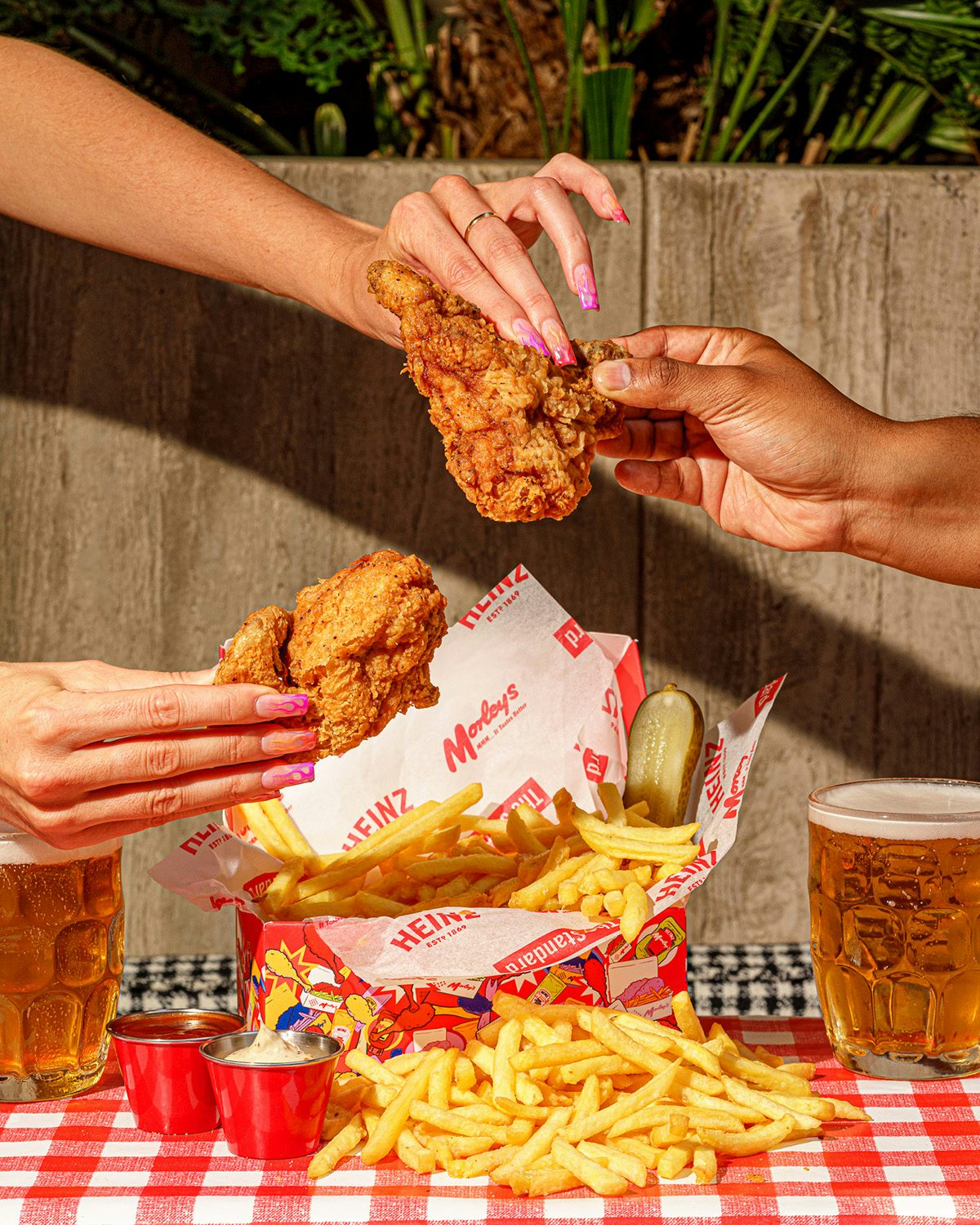 Photograph showing two people holding onto the same piece of fried chicken, leaning over a tablecloth covered in chips, pots of sauce, a box of chicken, and two glasses of beer