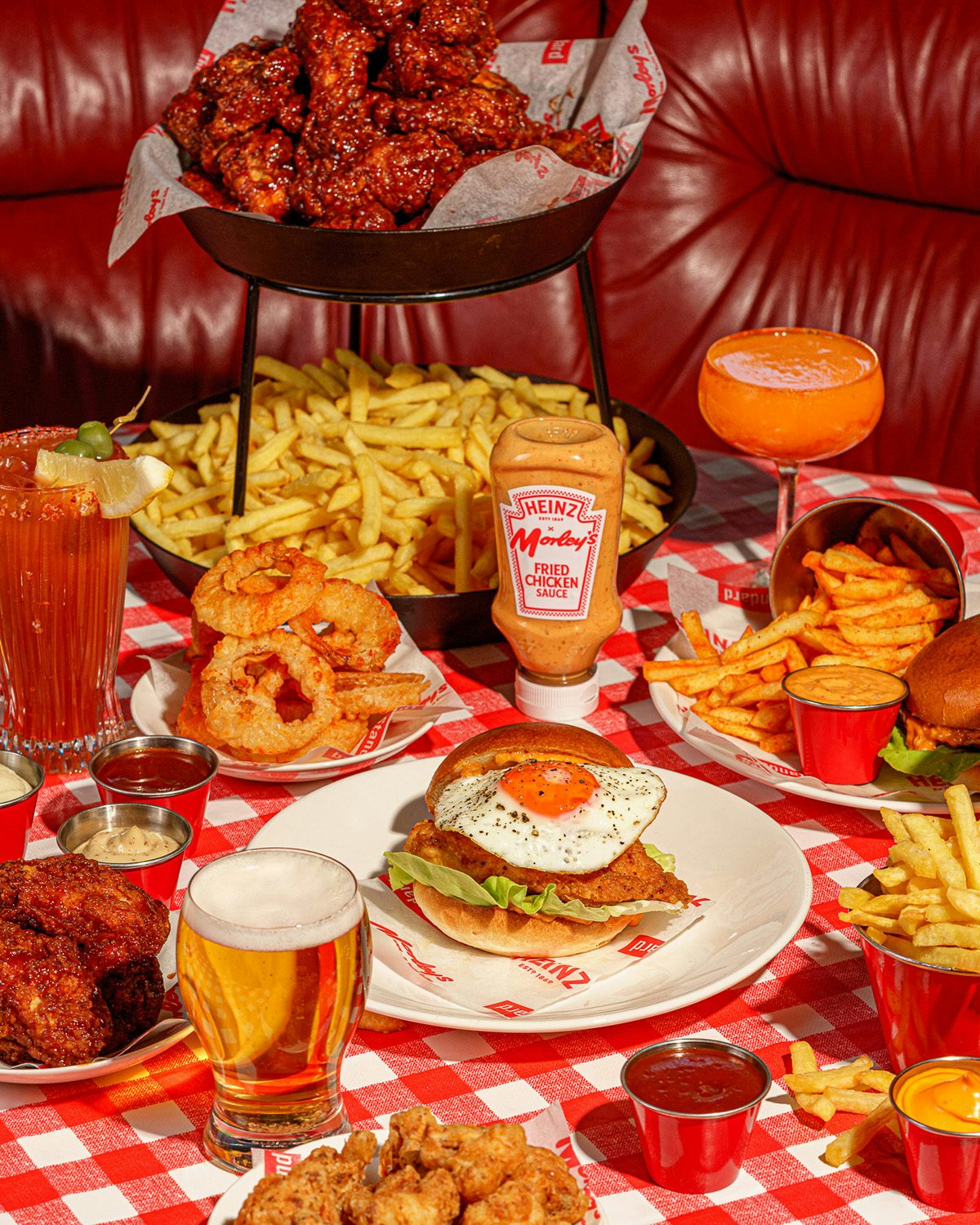 Photograph of a red gingham tablecloth covered in plates of food, including a chicken burger with an egg inside, onion rings, popcorn chicken, and chips, as well as orange cocktails and glasses of beer, as part of the Morley's and Heinz collaboration