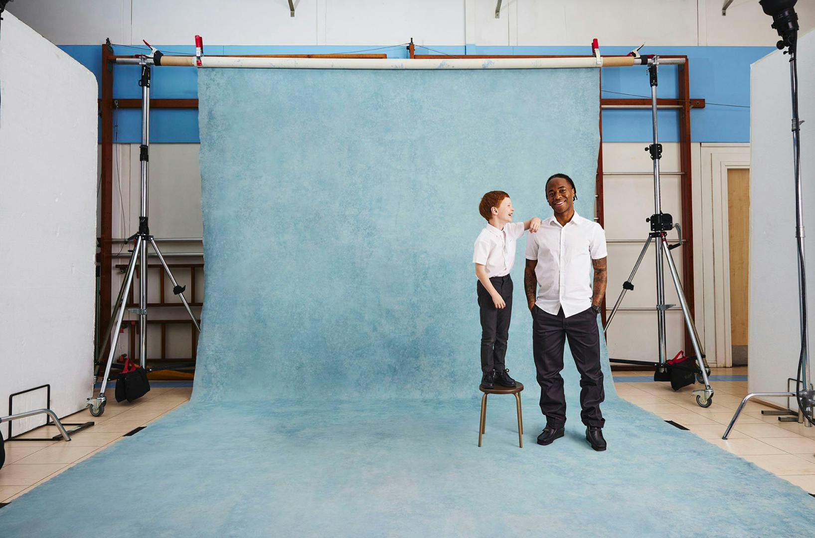 Photograph of Raheem Sterling wearing a white shirt and dark trousers with his hands in his pockets, with a school boy in uniform standing on a stool and leaning his arm on Sterling's shoulder. Both are standing on a blue studio backdrop