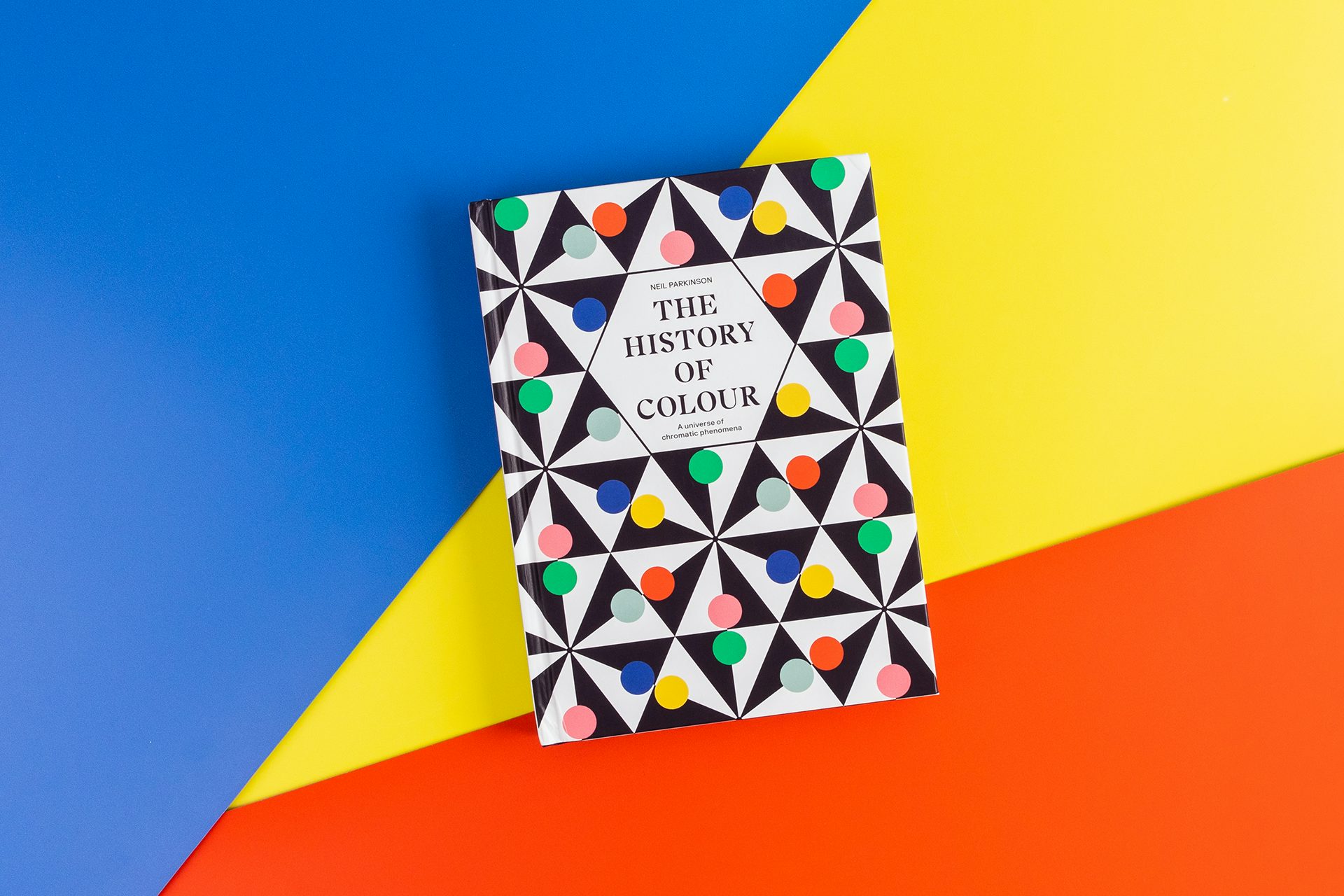 Image shows the cover of The History of Colour book, featuring a geometric and polka dot cover, arranged on a blue, yellow and red geometric background