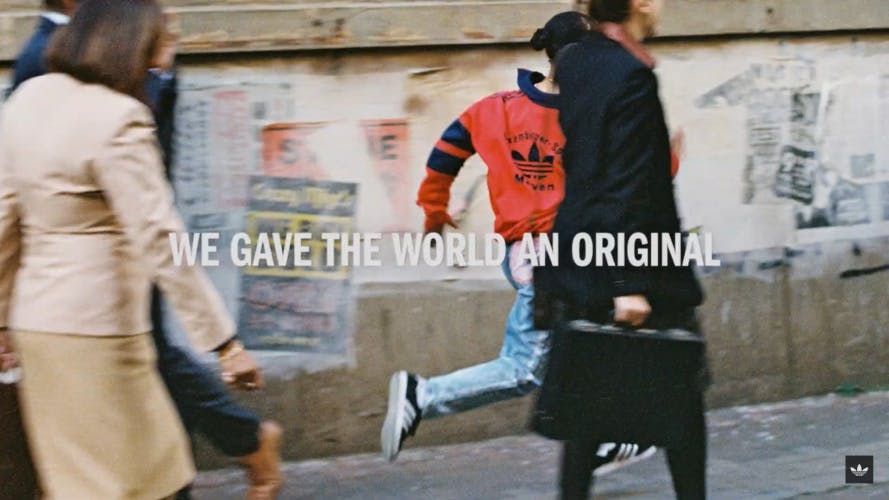 Still image from a new adidas Originals brand campaign showing an image of a person wearing jeans, a red adidas jacket, and black adidas trainers running next to a wall through a busy street, with the line 'We gave the world an original' laid over the image in white serif capitals