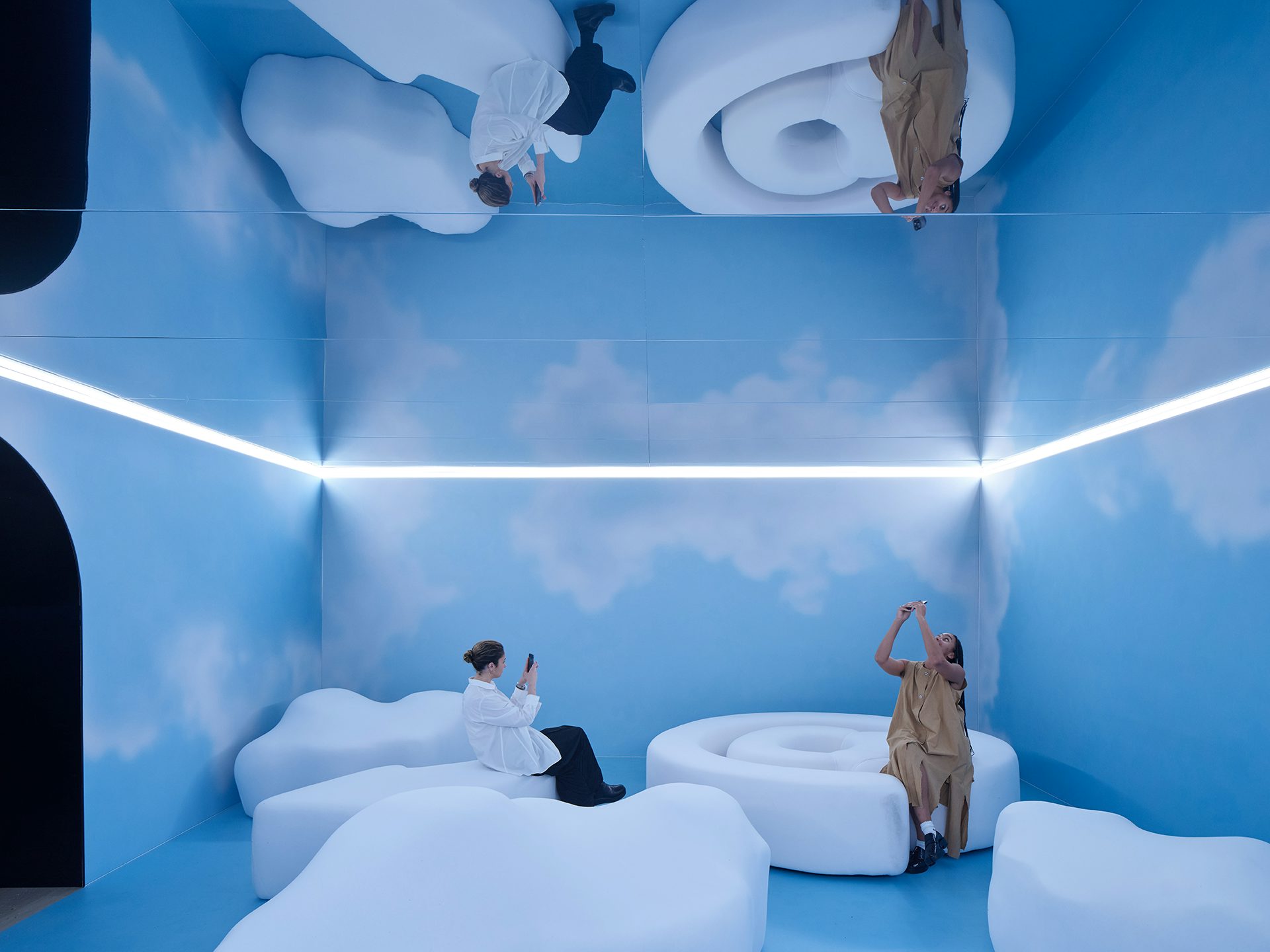 Photo shows two people taking photos in a room at Mailchimp's Email is Dead exhibition at the Design Museum, featuring large white pillowy seating, blue walls decorated with clouds, and a mirrored ceiling
