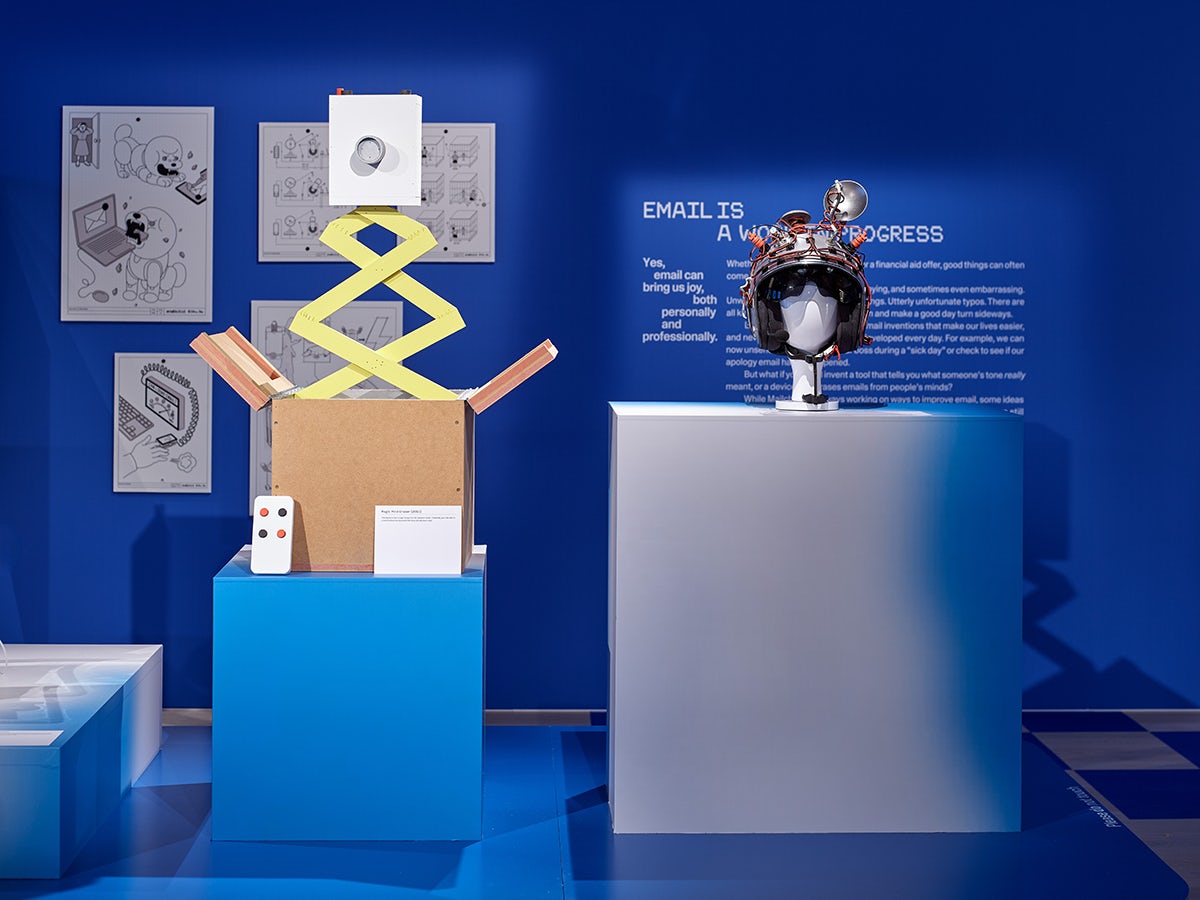 Photo shows an install at Mailchimp's Email is Dead exhibition at the Design Museum, featuring a large white cube with a helmet-like device on top, and a blue cube with a jack-in-the-box style device on top.