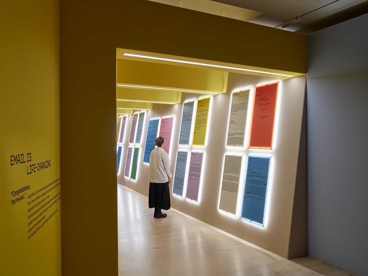 Photo shows a person wearing a beige coat and a dark skirt looking at an install at Mailchimp's Email is Dead exhibition at the Design Museum, featuring grids of four multicoloured plaques arranged in a row along a tunnel