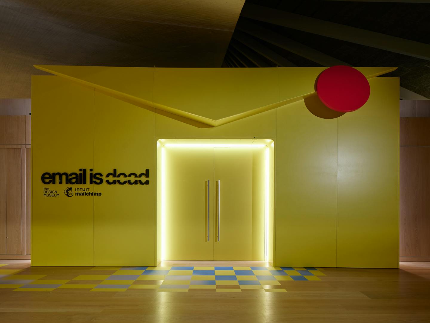 Photo of the entrace to Mailchimp's Email is Dead exhibition at the Design Museum, featuring a doorway in the middle of a large yellow entrance in the shape of an envelope and the exhibition title written on the left of the envelope