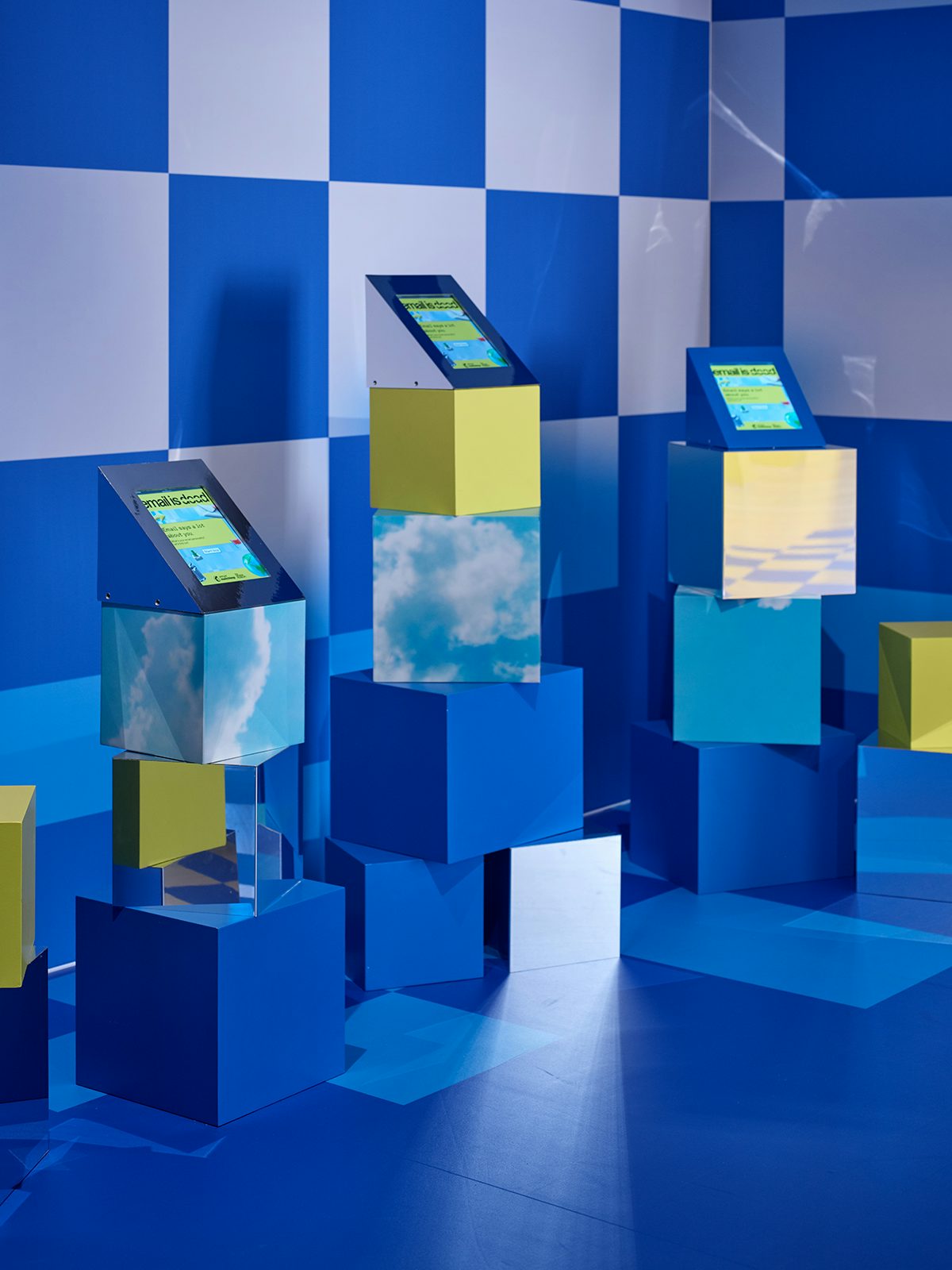 Image shows an install at Mailchimp's Email is Dead exhibition at the Design Museum, featuring blue and yellow blocks stacked unevenly with three touch screen devices arranged on top of each stack
