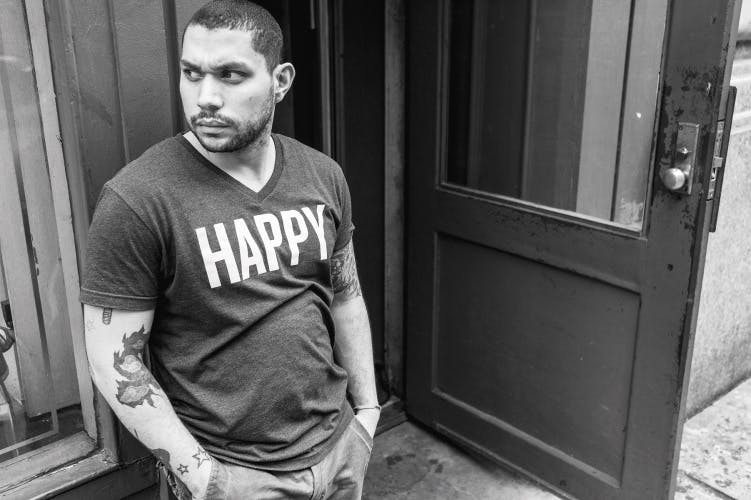 Black and white photograph from Selected Writings by Richard Kalvar showing a person with very cropped hair, a short beard and tattoos wearing a t-shirt that reads 'happy'