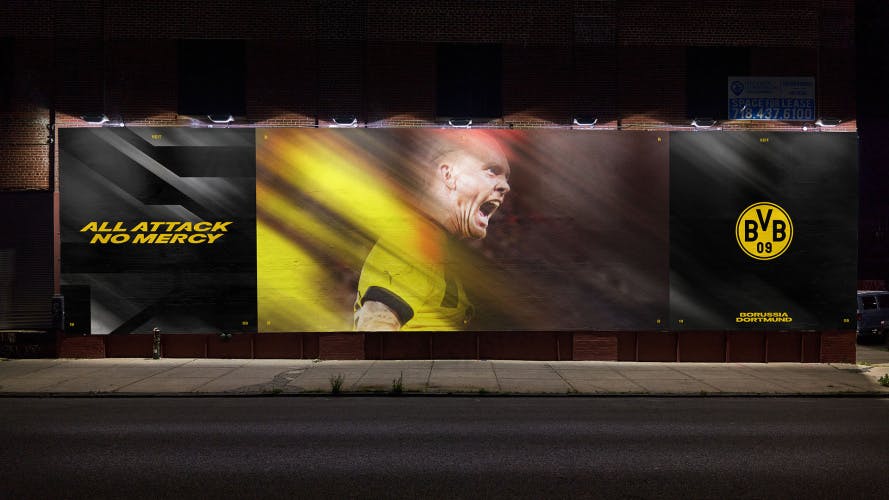 Image shows BVB Borussia Dortmund's branding on a wide outdoor display advertisement, which reads 'All attack no mercy' in yellow slanted lettering, the club's badge, and a blurred image of a player in the middle