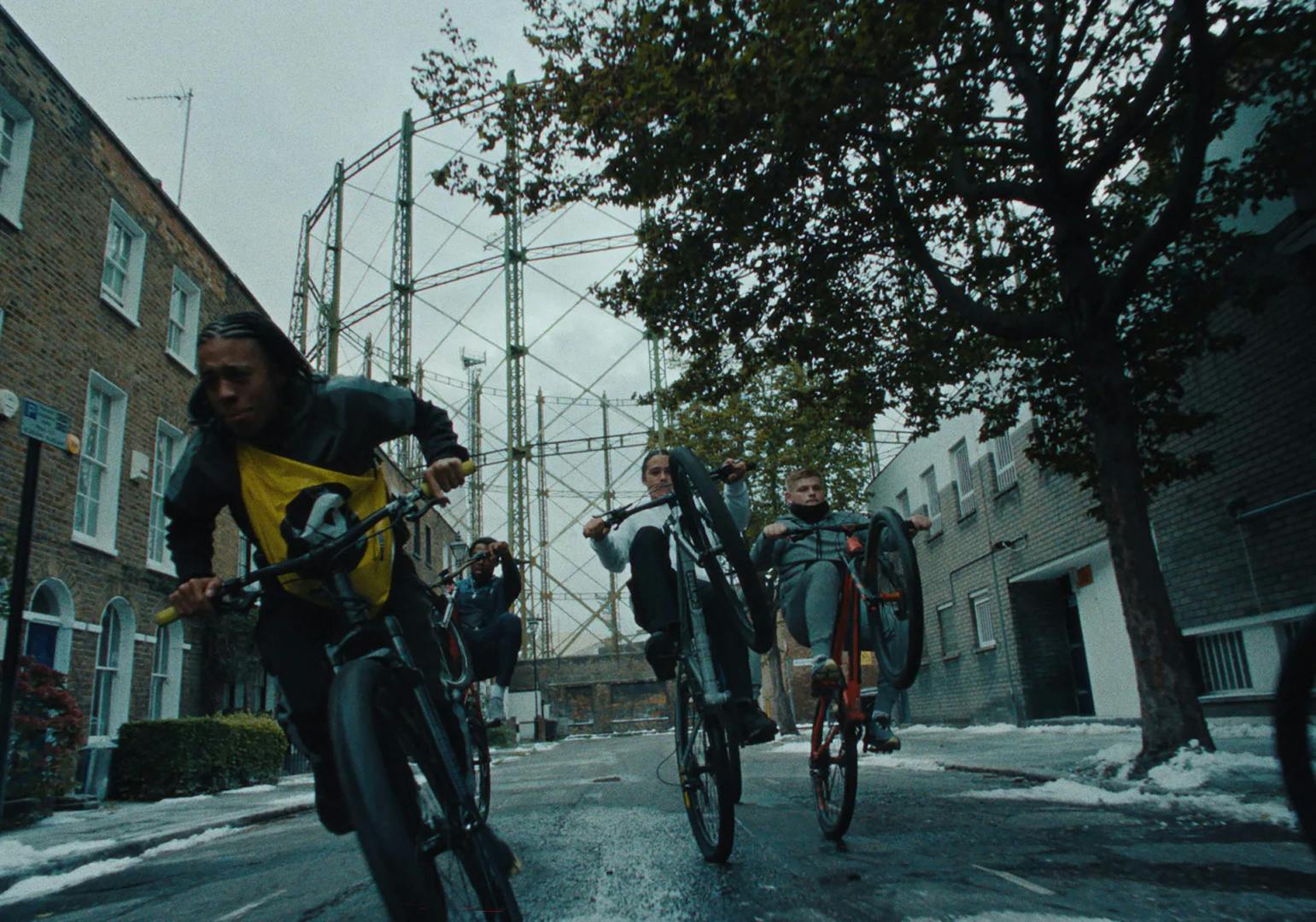 Still from JD Sports Christmas 2023 ad showing three young people doing bike wheelies down a residential street with a gasholder in the background
