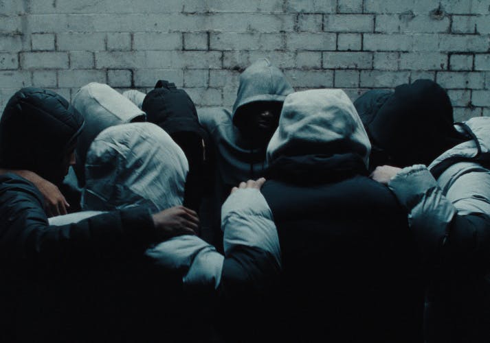 Still from JD Sports Christmas 2023 ad showing a crowd of young people with hoods pulled over their heads forming a circle with their arms around one another