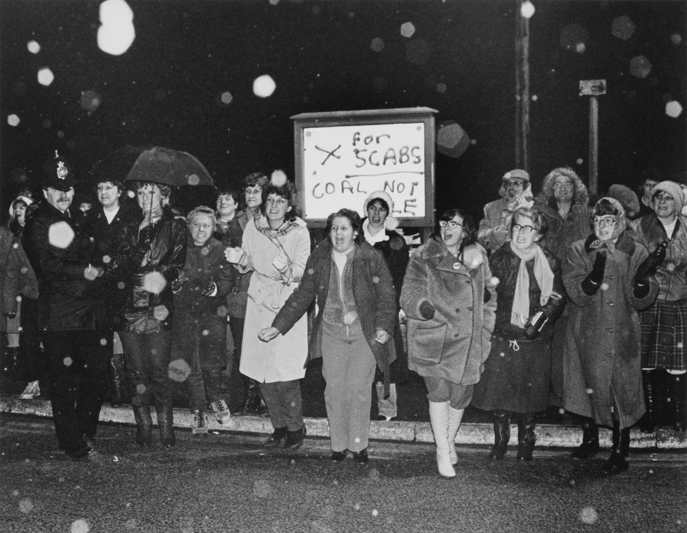 Black and white photograph of a line of women protestors wearing winter coats and carrying a sign that reads 'X for scabs, coal not dole'
