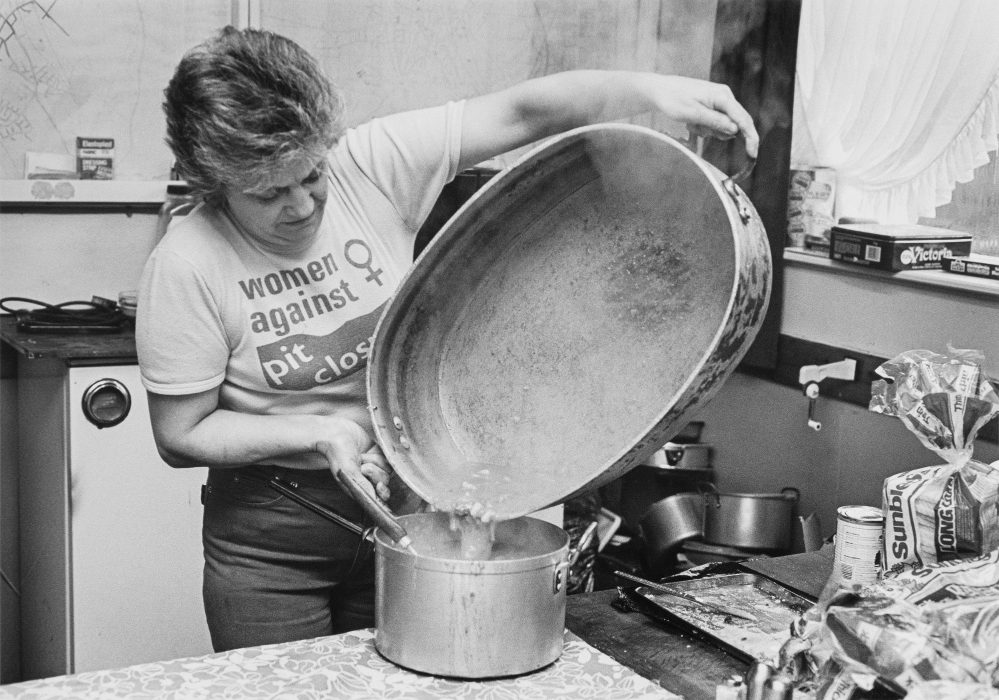 Black and white photograph of a woman pouring food from a very large metallic pot into a smaller pot, wearing jeans and a t-shirt that reads 'women against pit closures'