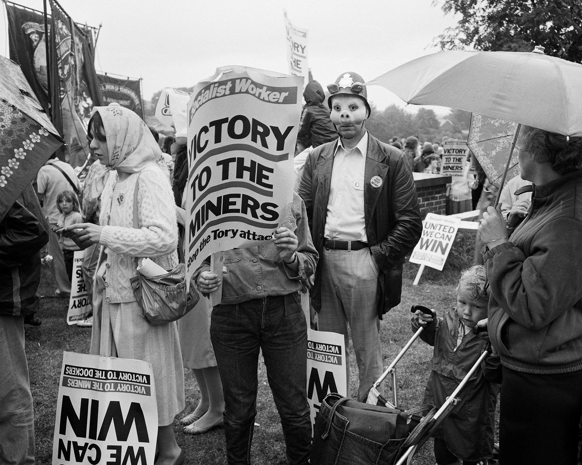 Black and white photograph of protestors holding signs that read 'Victory to the miners' and 'United we can win'. At the centre of the image is a person wearing a pig mask and a police helmet with pig ears protruding from it
