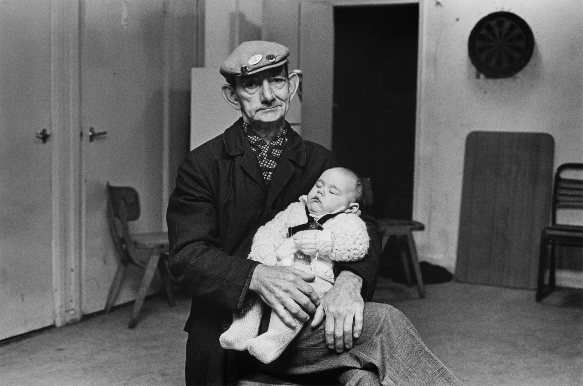Black and white photograph of a retired pit deputy wearing a hat and dark coat holding a three-month-old baby sleeping on his lap