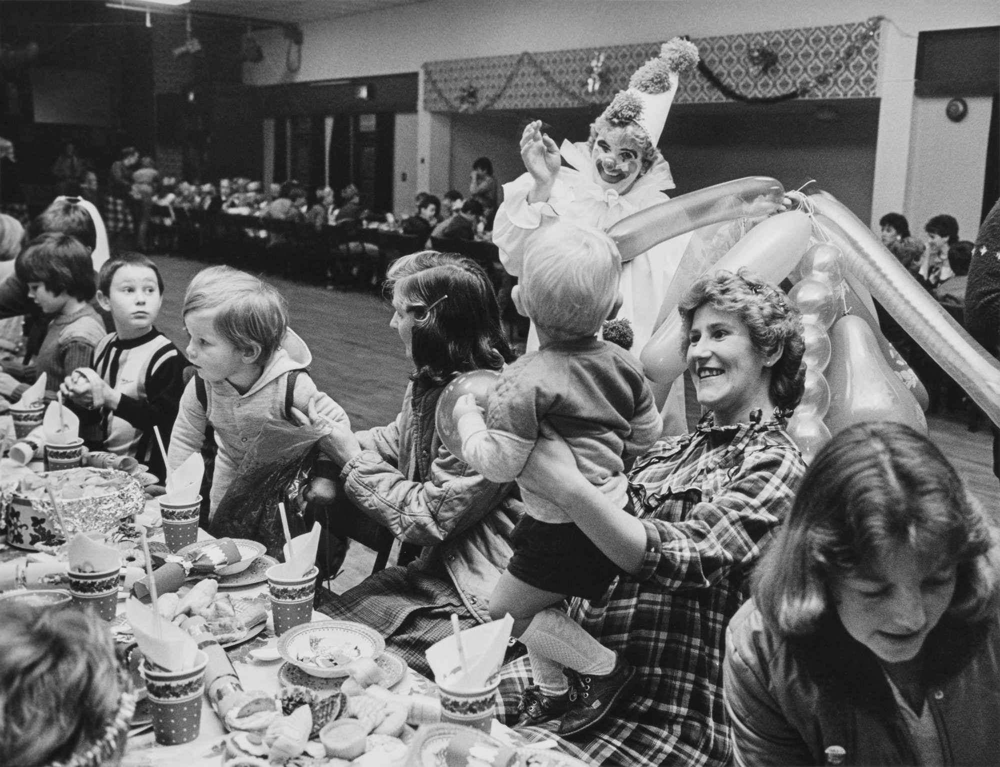 Black and white photograph showing parents and children of striking miners eating party food from long tables inside a hall