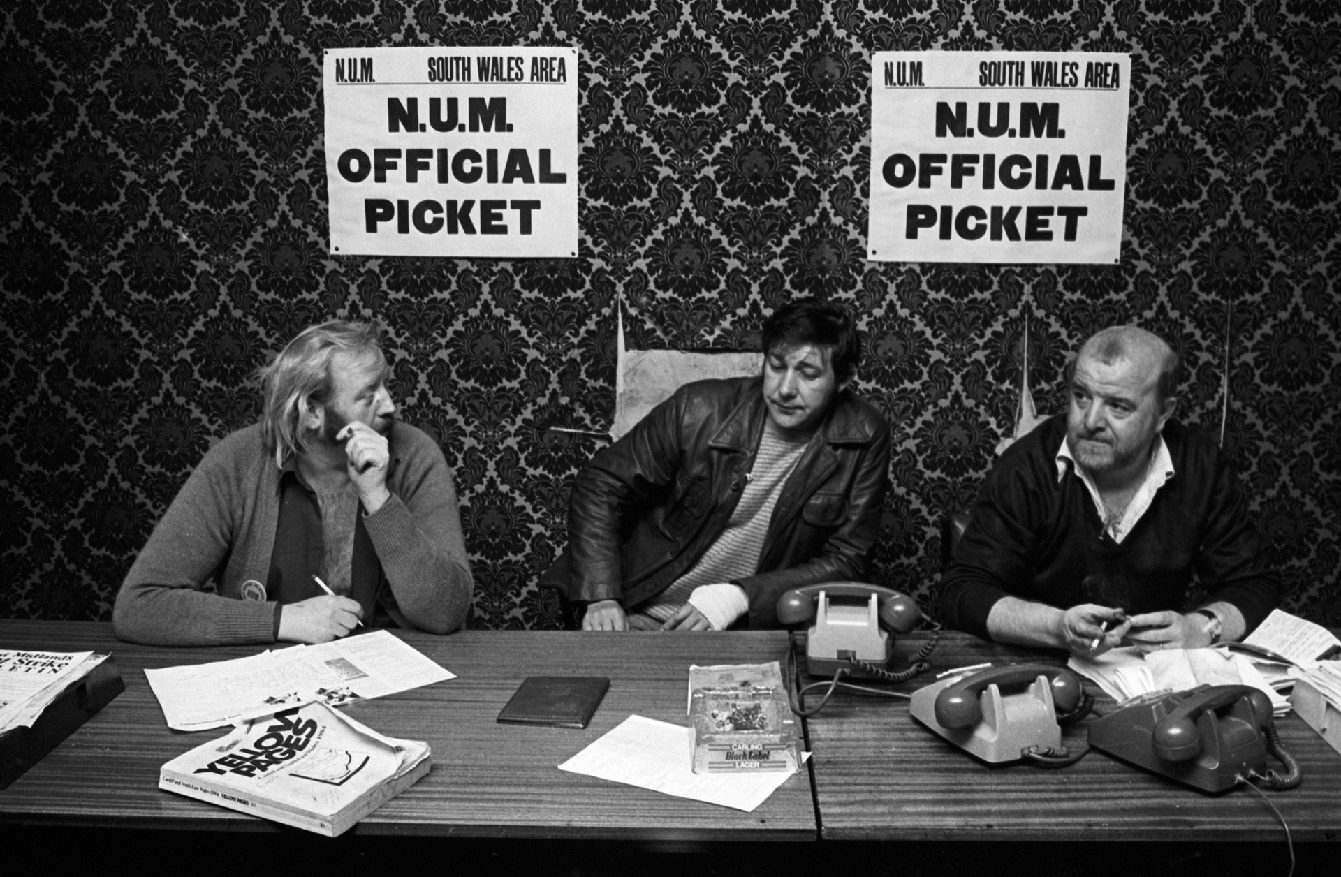 Black and white photograph of three union officials sat at a desk covered in phones and a Yellow Pages book, with two signs that read 'N.U.M Official Picket' stuck to a patterned wall behind them