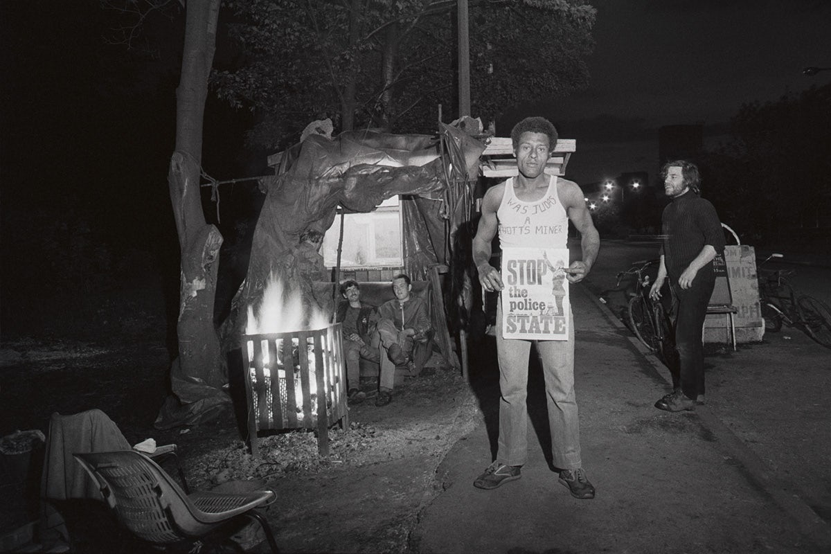 Black and white photograph of a person holding a sign that reads 'Stop the police state' and wearing a light vest that reads 'Was Judas a Notts Miner?'. In the background, two people are seat inside a DIY structure with a burning crib in front of them