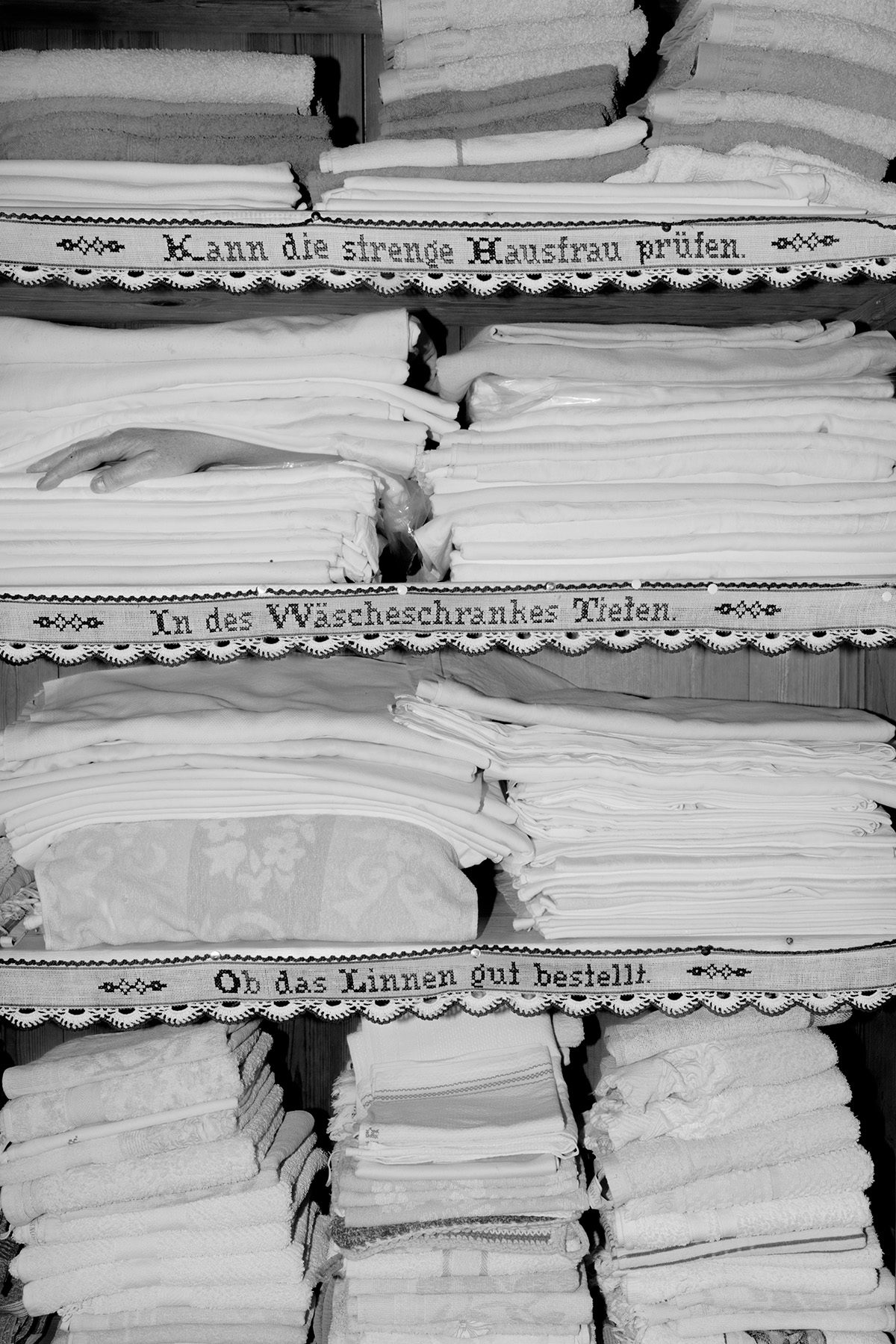 Black and white photograph from Plexus by Elena Helfrecht showing piles of linens, which have been folded on shelves that are edged with German phrases. A hand is nestled between the piles