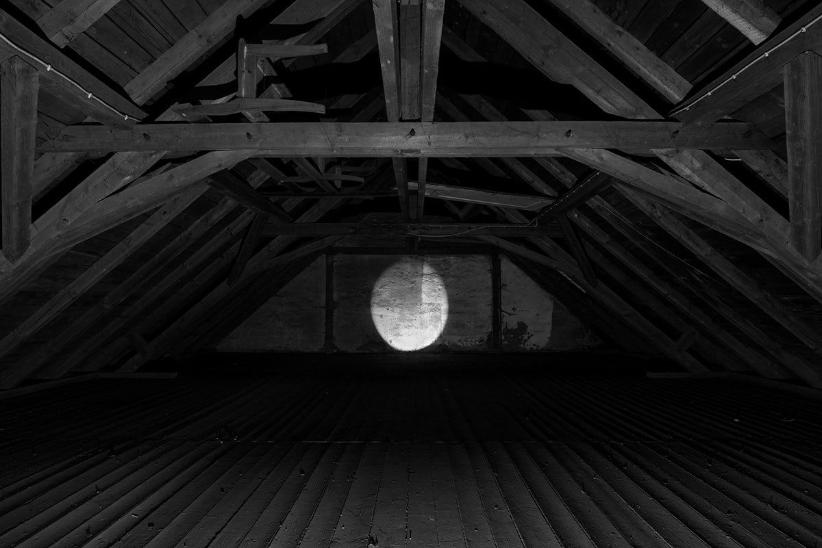 Black and white photograph from Plexus by Elena Helfrecht showing a dark attic space with a moon-shaped light beam on the wall