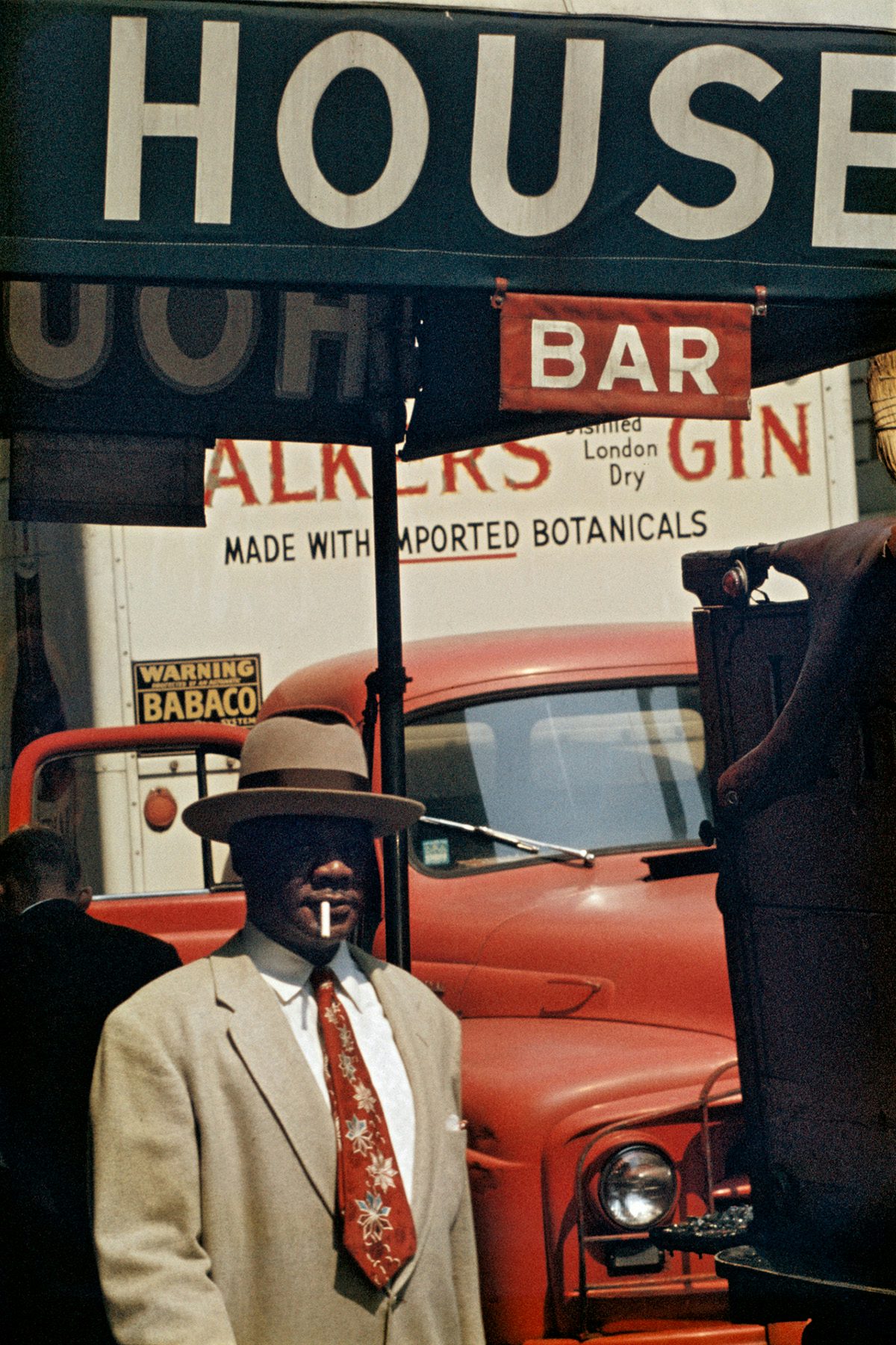 Photograph by Saul Leiter showing a man standing in front of a red vehicle wearing a camel coloured hat and suit with a cigarette in his mouth