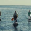 Still image from an advert for Avios showing a row of 7 people riding efoil boards over a body of water while carrying shopping bags