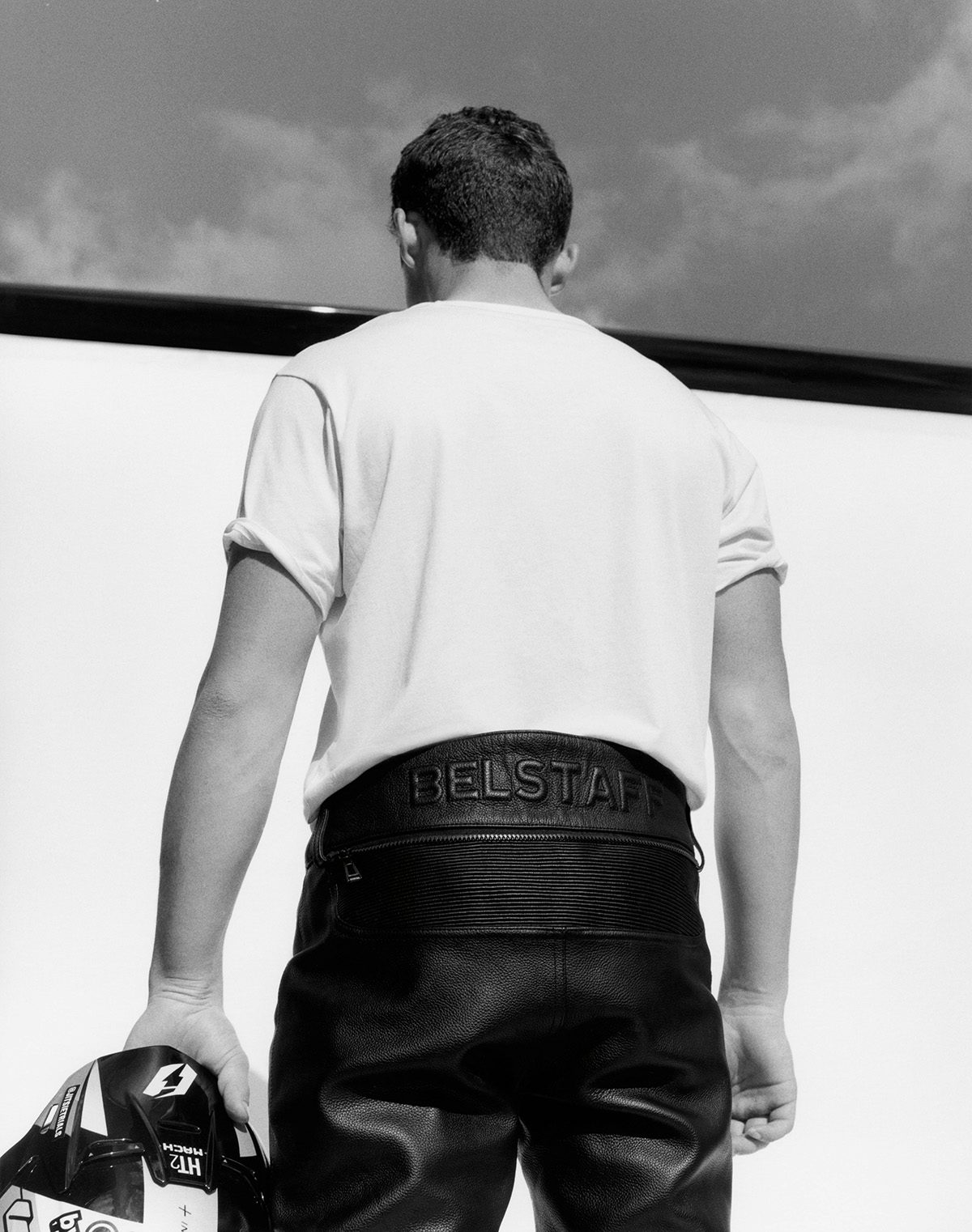 Black and white photo of a person stood with their back to the camera wearing leather trousers and white t-shirt taken from Belstaff centenary book